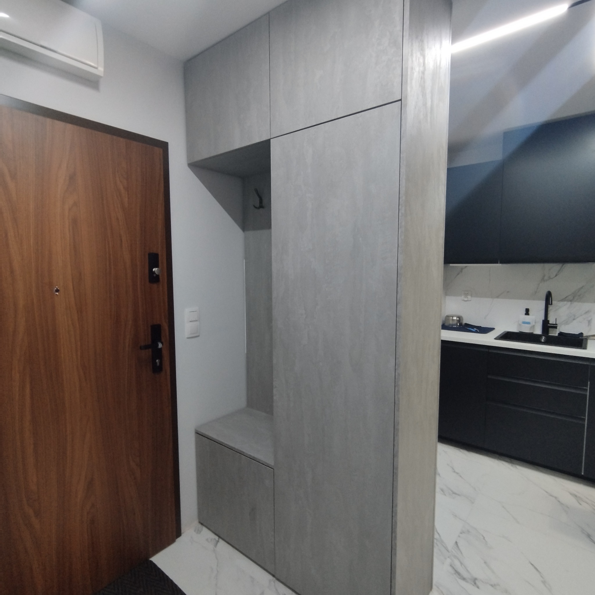 Szara zabudowa przedpokoju z siedziskiem i wieszakami, widok na kuchnię z czarnymi szafkami i marmurowym blatem.