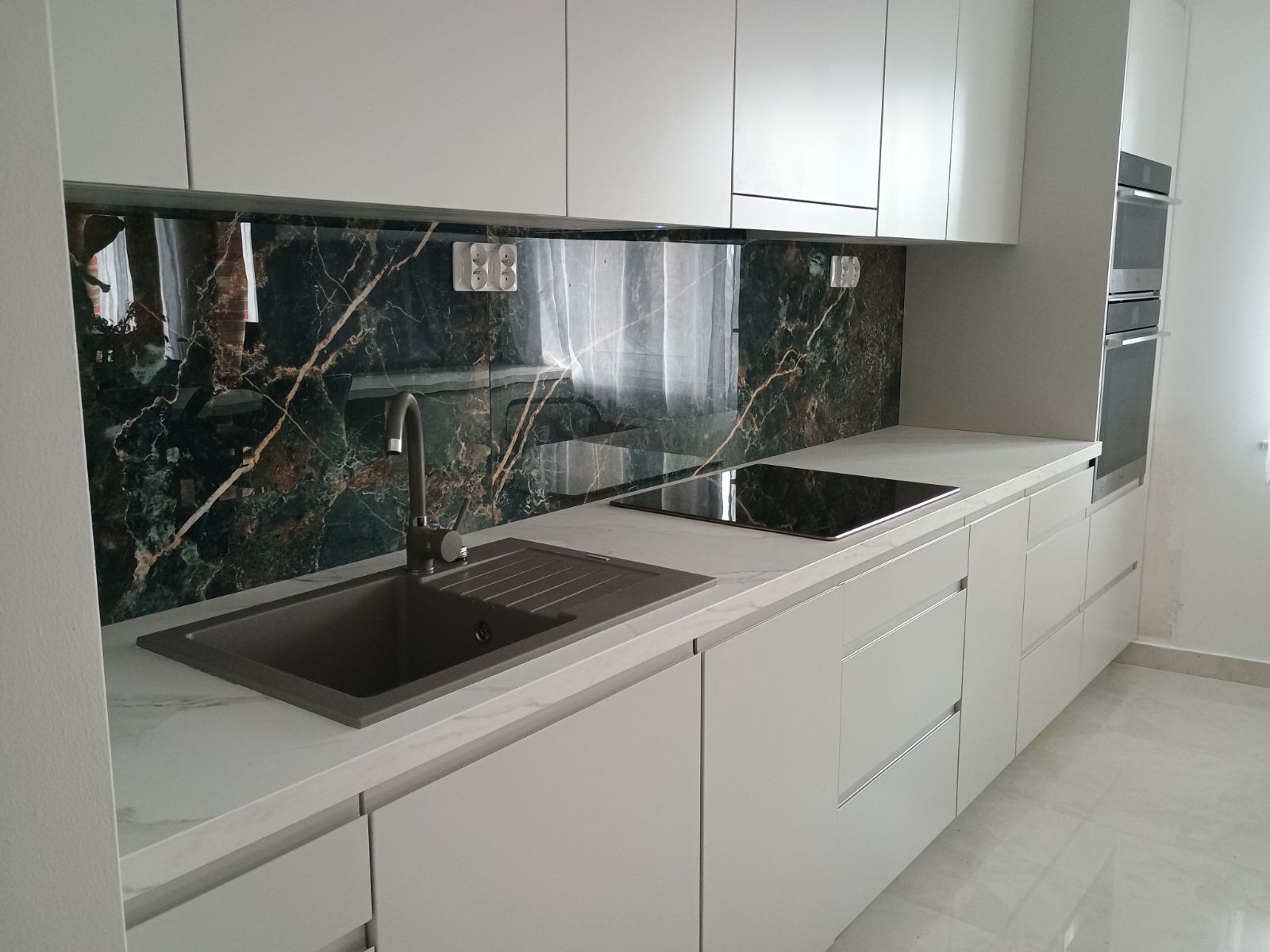 Nowoczesna kuchnia z białymi szafkami, blatem imitującym marmur i panelem szklanym nad blatem z motywem ciemnego marmuru z jasnymi żyłami. Widoczny zlew w kolorze grafitowym i płyta indukcyjna.