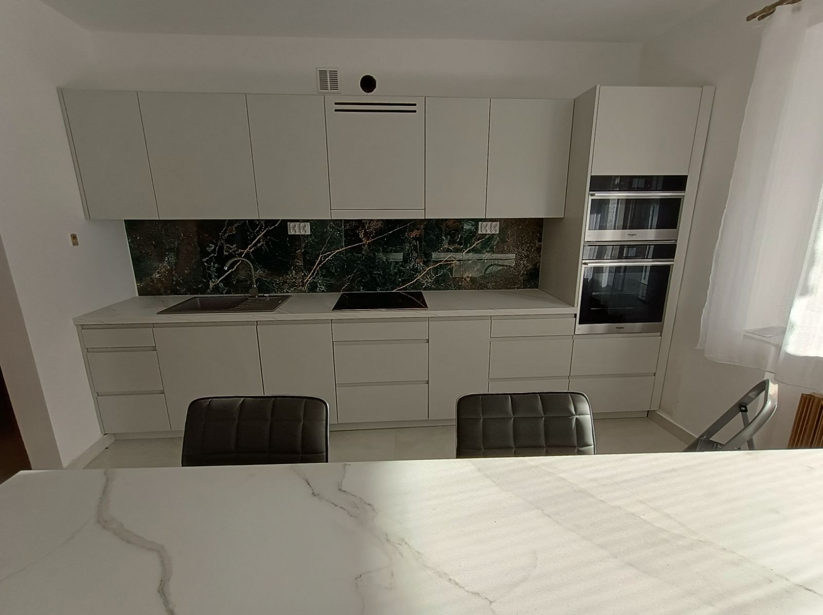 Nowoczesna kuchnia na wymiar z białymi frontami, blatem imitującym marmur i zielonym panelem nad blatem, widok zza stołu z marmurowym blatem.