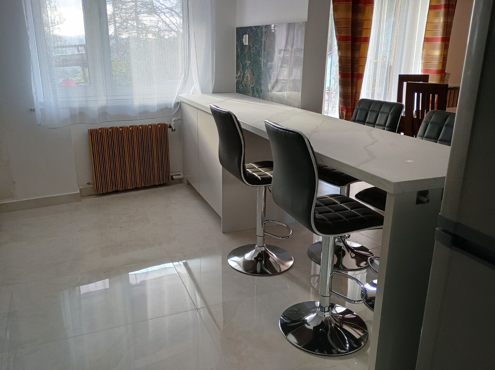 Wysoki, biały blat z marmurowym wzorem, wsparty na szafkach, z trzema nowoczesnymi, czarnymi hokerami na chromowanych nogach. W tle okno z firanką i grzejnik żebrowy.