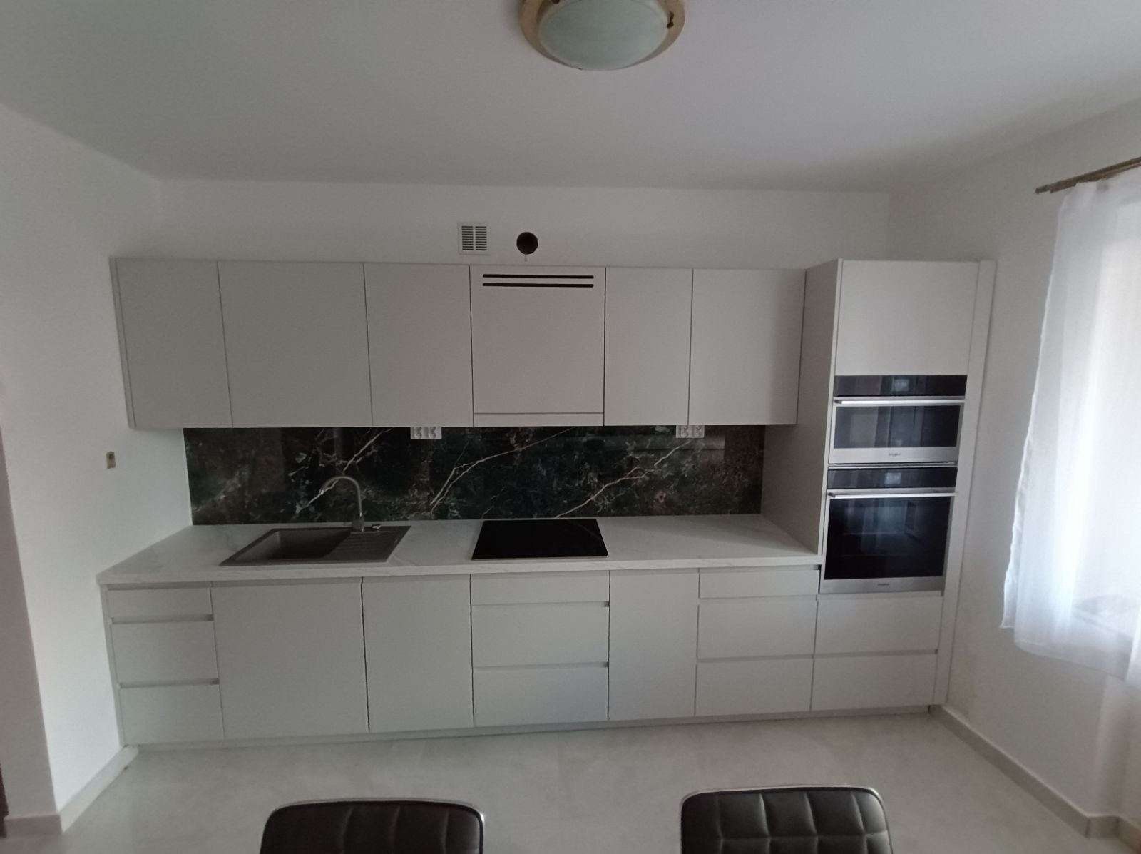 Jasna, nowoczesna zabudowa kuchenna z frontami bez uchwytów, blatem w kolorze marmuru i ciemnozielonym panelem nad blatem, z wbudowanym piekarnikiem i płytą grzewczą.