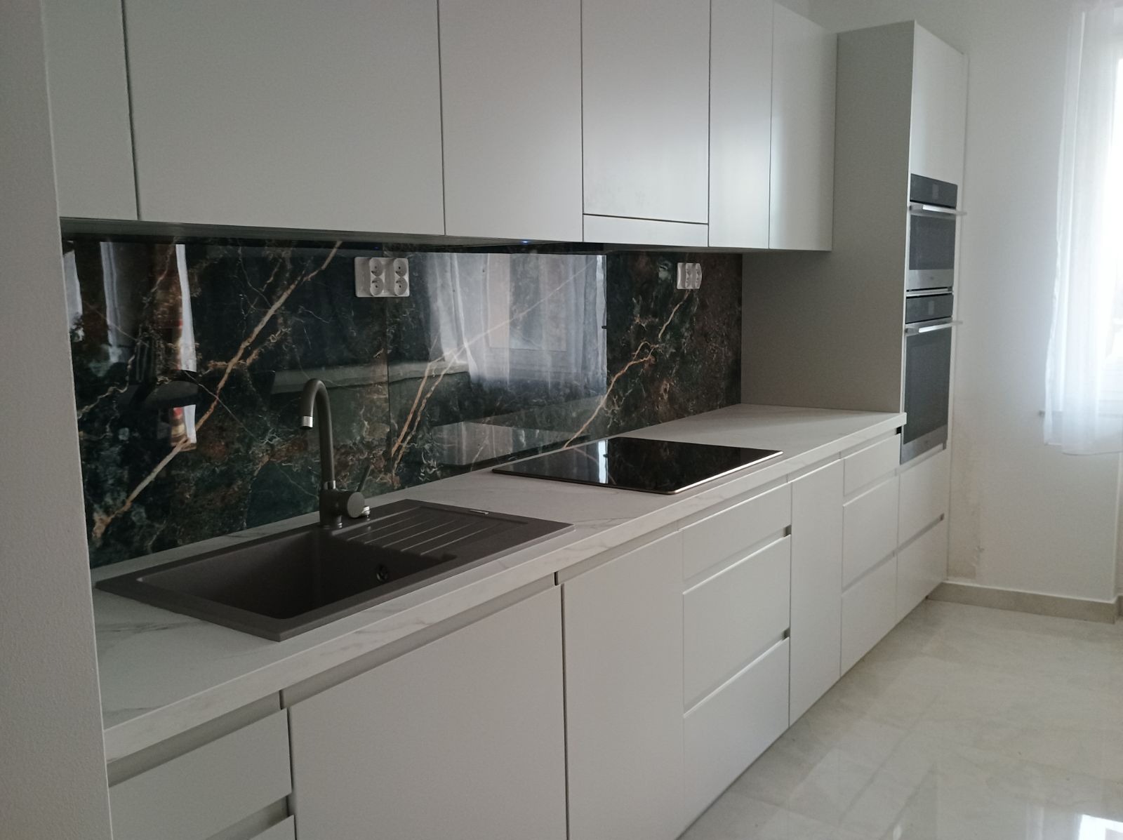 Nowoczesna kuchnia z białymi, matowymi frontami, blatem imitującym marmur i czarnym panelem nadblatowym z motywem marmuru. W tle wbudowane piekarniki.