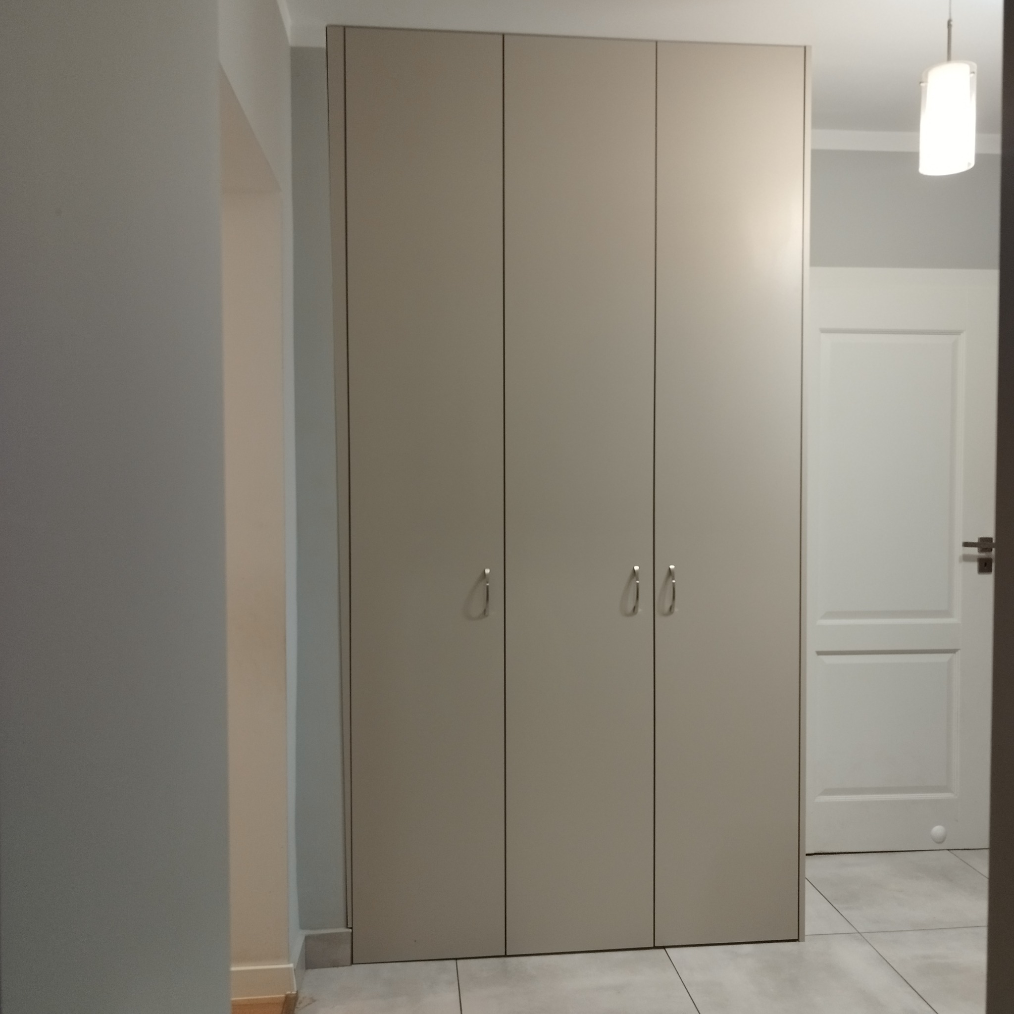 Czterodrzwiowa, beżowa szafa sięgająca sufitu, z minimalistycznymi, podłużnymi uchwytami, wkomponowana w jasne wnętrze z białą podłogą i drzwiami.