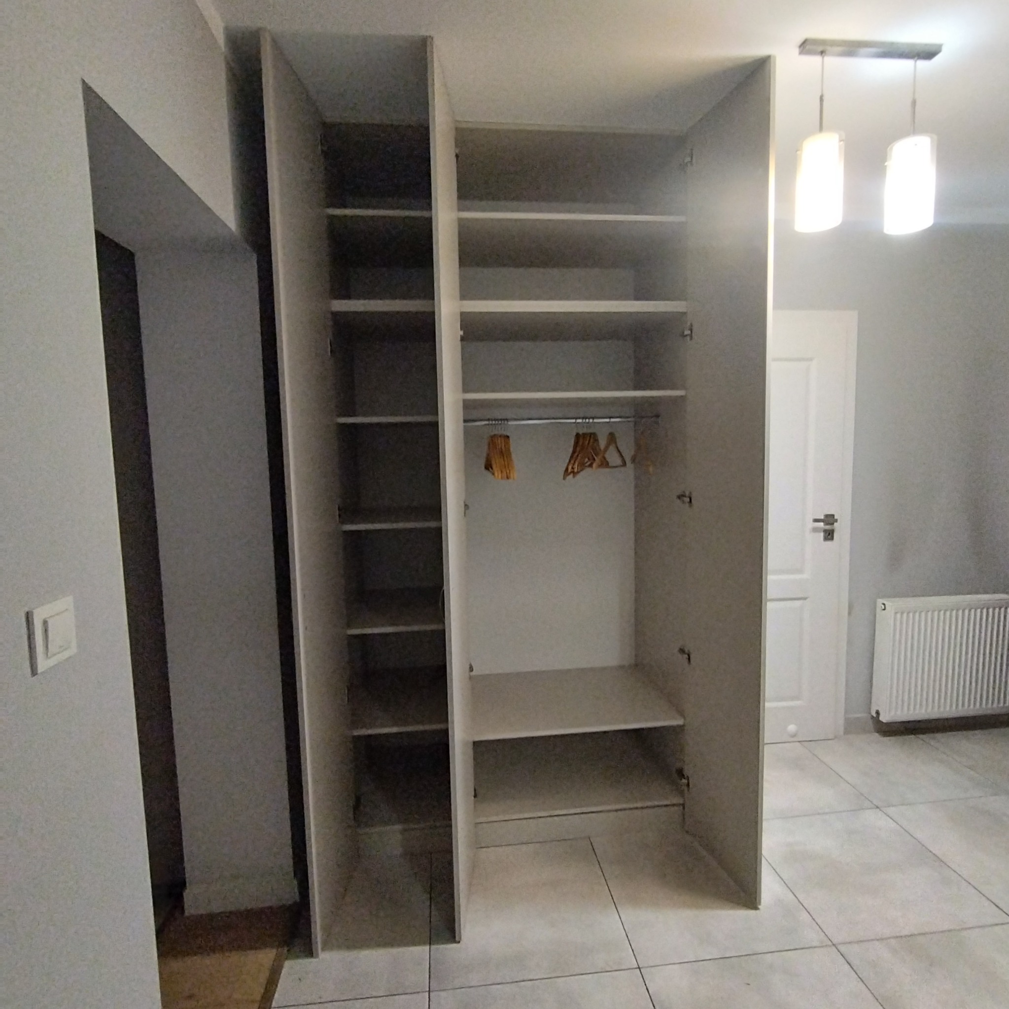 Wnętrze szafy z półkami i drążkiem na wieszaki, widoczne wieszaki bez ubrań, oświetlenie sufitowe w tle, jasna kolorystyka wnętrza.