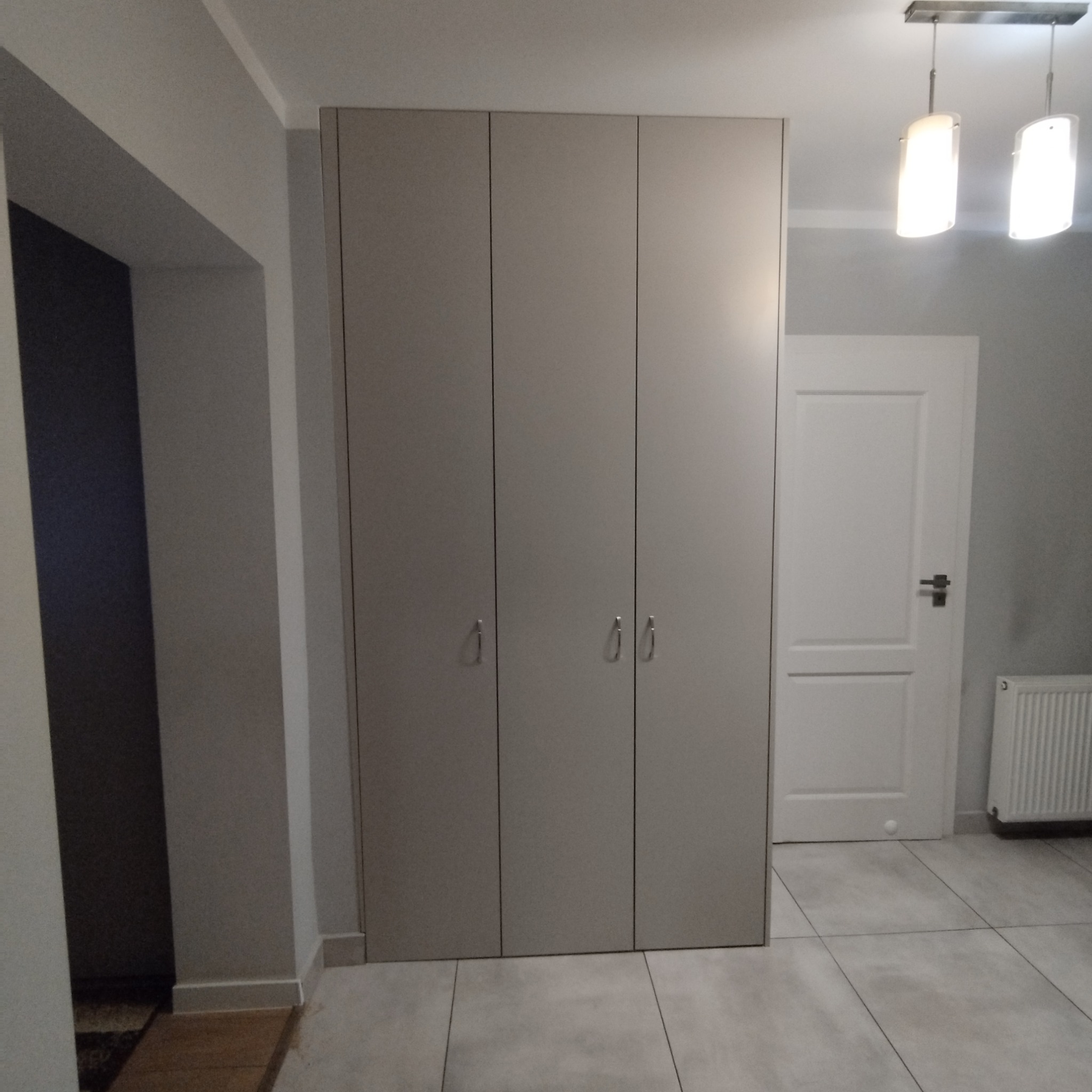 Trzydrzwiowa, beżowa szafa wnękowa z metalowymi uchwytami na tle białej ściany, drzwi i grzejnika, oświetlona lampą sufitową