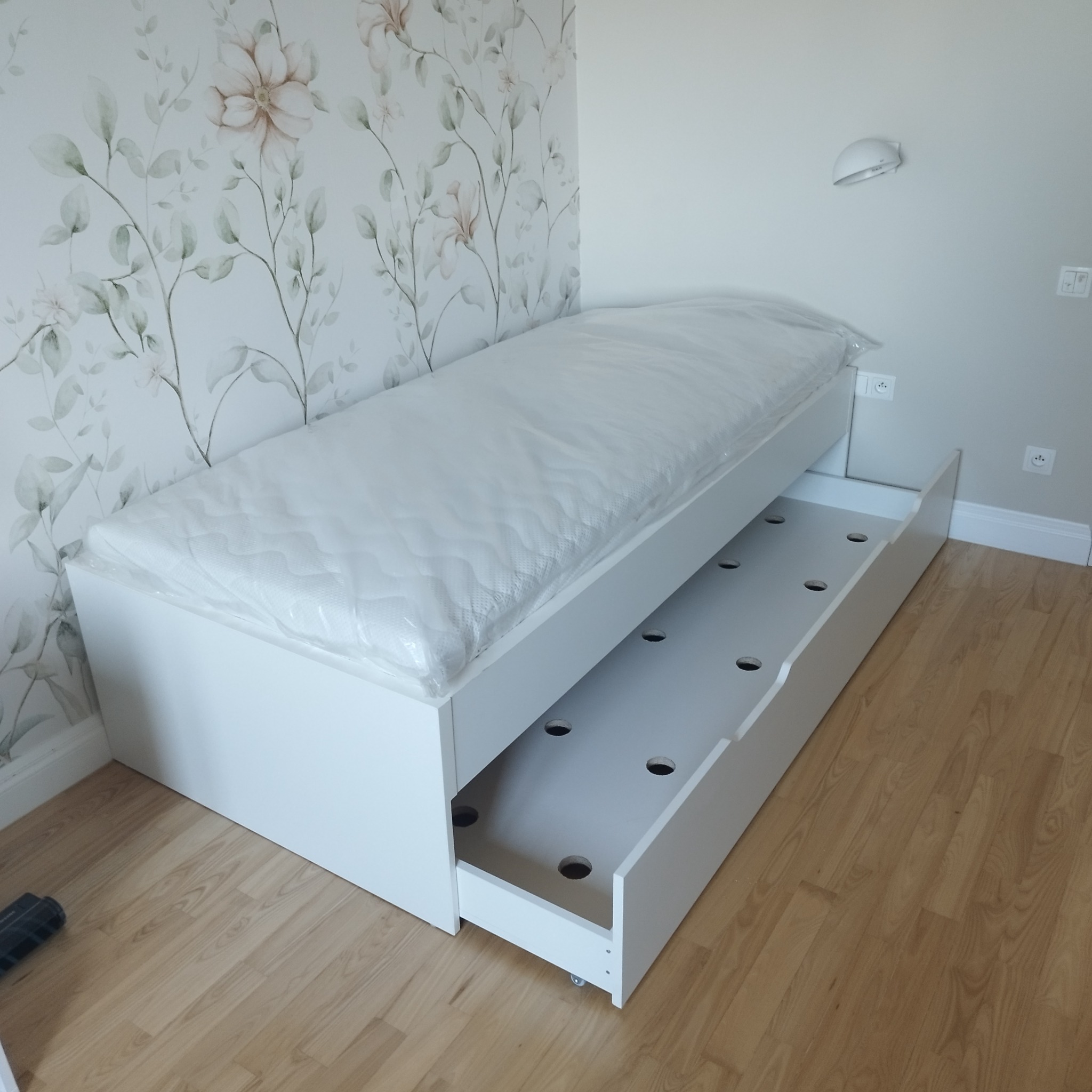 Białe łóżko z wysuwanym dodatkowym spaniem, materac w folii, na podłodze panele, ściana z tapetą w kwiaty.