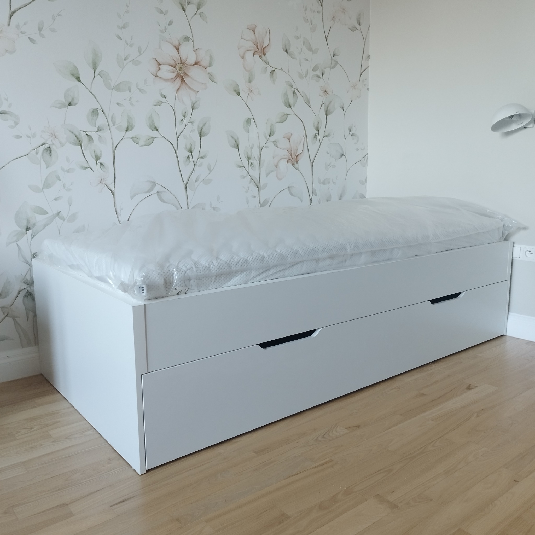 Białe łóżko z szufladami na tle ściany z motywem kwiatowym. Materac owinięty folią ochronną.