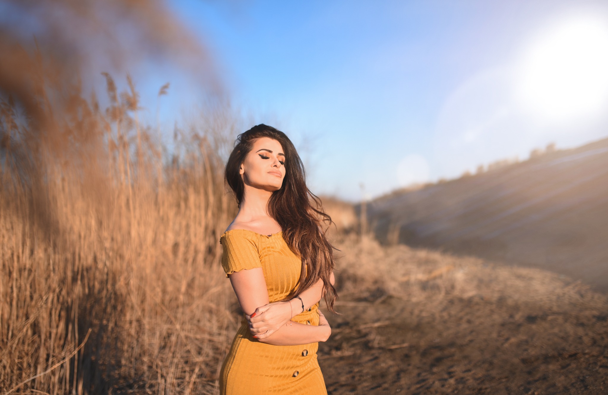 Kobieta w musztardowej sukience pozuje w plenerze na tle wysokich traw i rozświetlonego słońcem nieba.