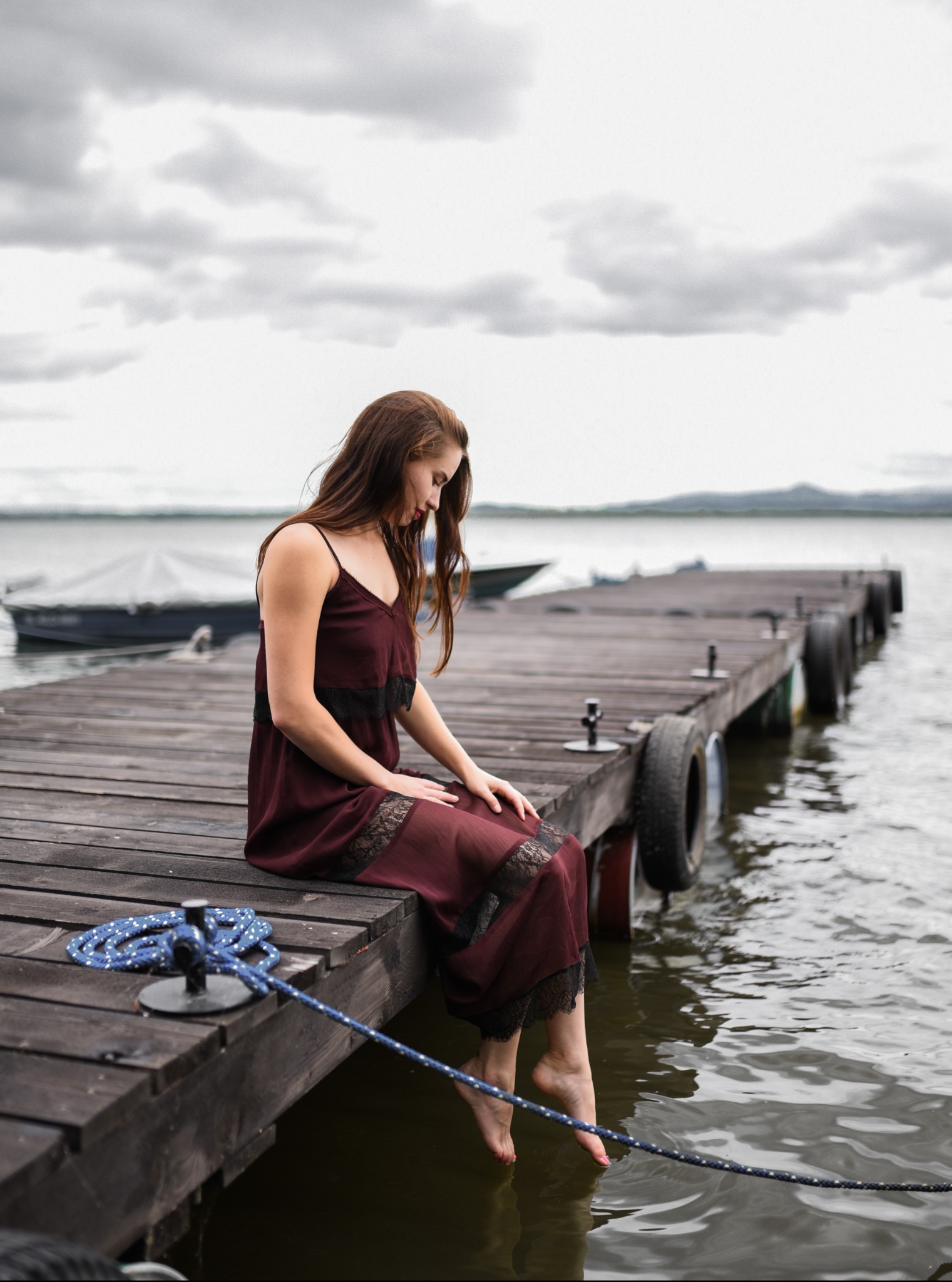 Kobieta w bordowej sukience z koronką siedzi na drewnianym pomoście nad jeziorem, stopy zanurzone w wodzie, w tle łodzie i pochmurne niebo.
