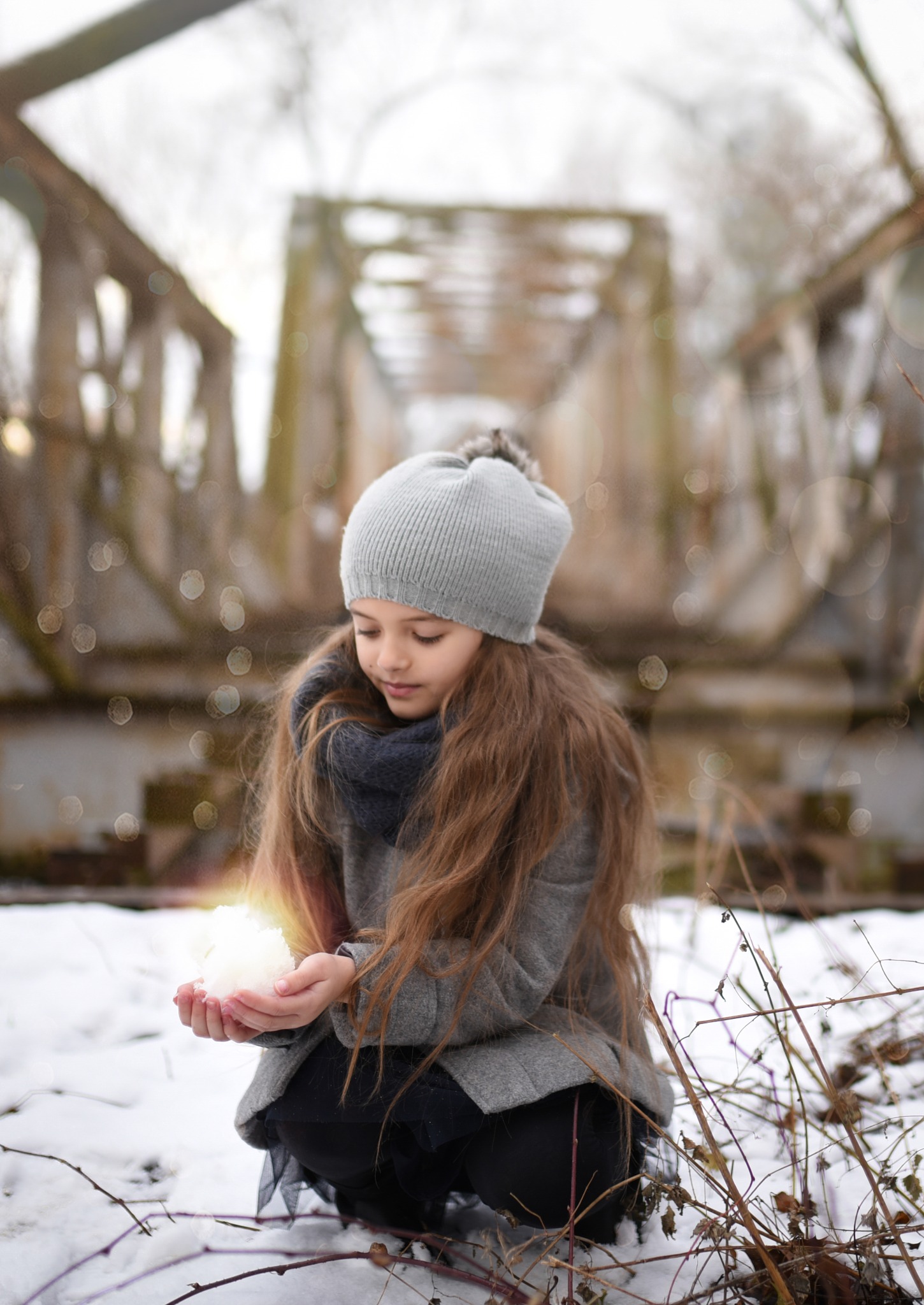 Dziewczynka w szarym płaszczu i czapce, kuca na śniegu przed starym metalowym mostem, trzymając w dłoniach kulę śniegu, która wydaje się świecić.