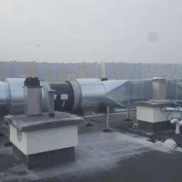 System kanałów wentylacyjnych na dachu budynku, widoczne elementy metalowe i plastikowe rury, w tle blok mieszkalny w porannej mgle.