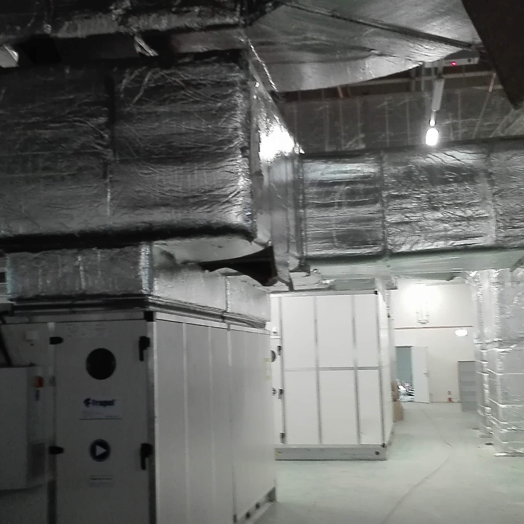 Widok na instalację wentylacyjną z izolowanymi kanałami pod sufitem w hali przemysłowej, widoczne białe centrale wentylacyjne z elementami sterującymi.