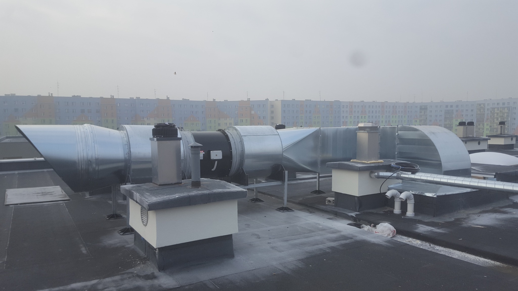 System kanałów wentylacyjnych na dachu budynku, widoczne elementy metalowe i plastikowe rury, w tle blok mieszkalny w porannej mgle.