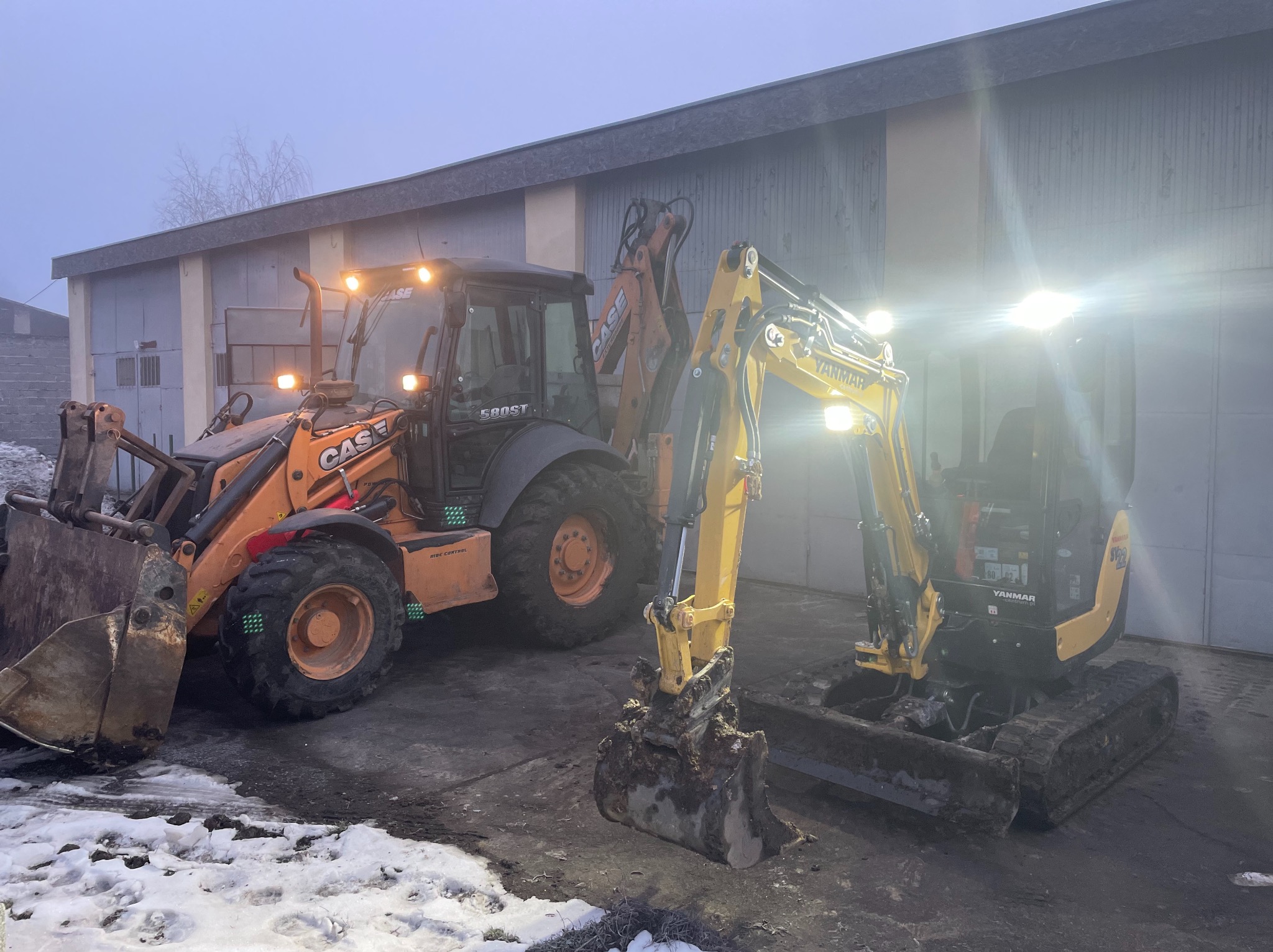 Pomarańczowa koparko-ładowarka Case 580ST i żółta mini koparka Yanmar SV22 ustawione obok siebie na placu budowy, w tle szary budynek magazynowy oświetlony światłami maszyn.