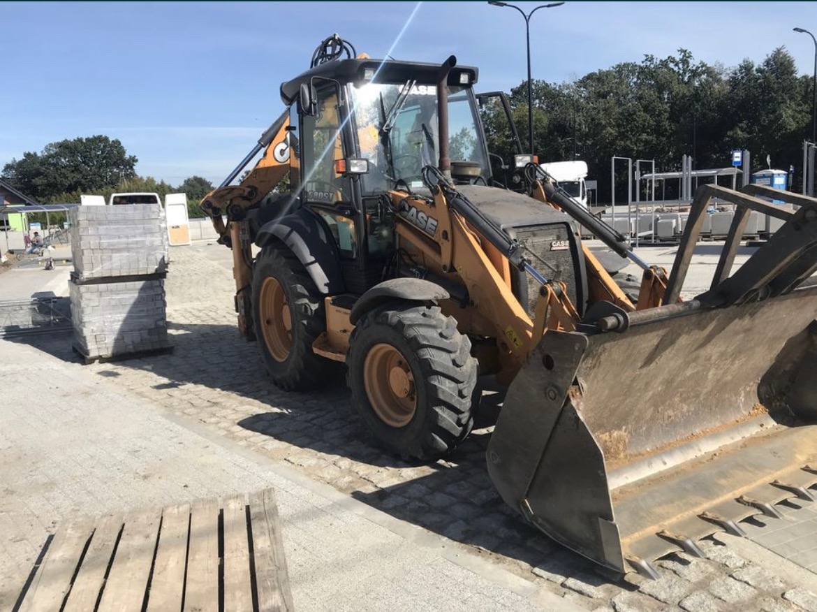 Żółto-czarna koparko-ładowarka marki CASE na kostce brukowej, obok paleta z kostką, w tle budynek w trakcie budowy i zaparkowany biały samochód dostawczy.