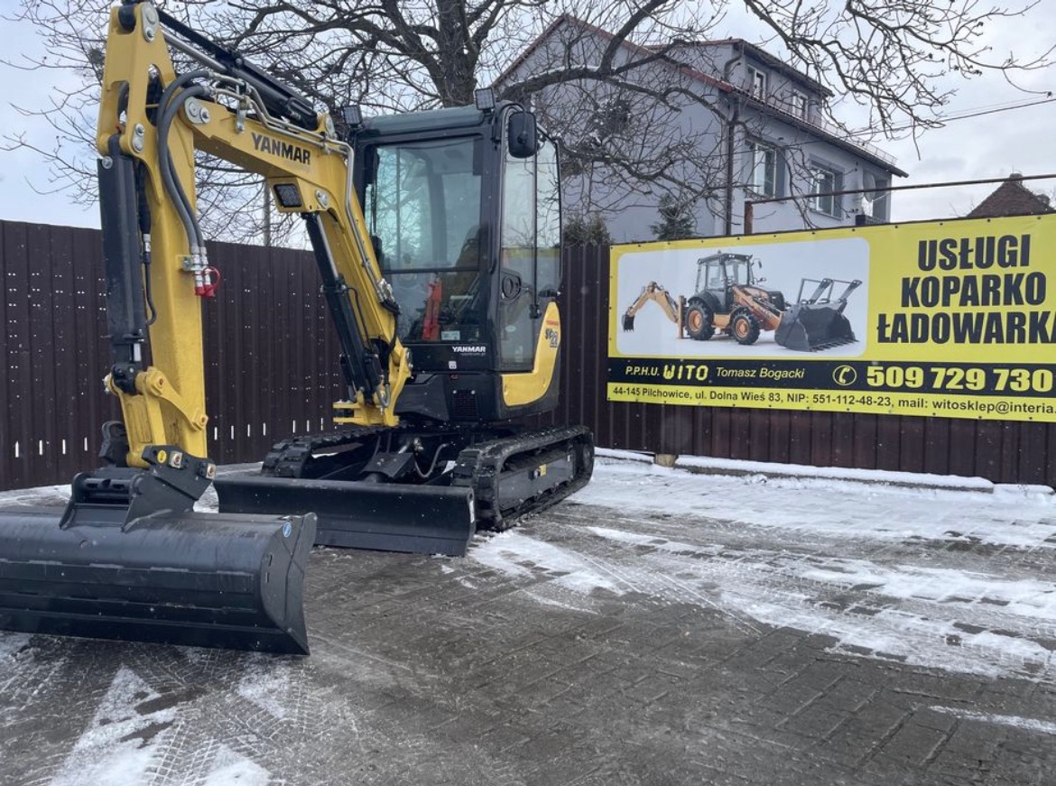 Żółta mini koparka Yanmar na tle ogrodzenia z banerem reklamowym usług koparko-ładowarką. Na ziemi leży śnieg.