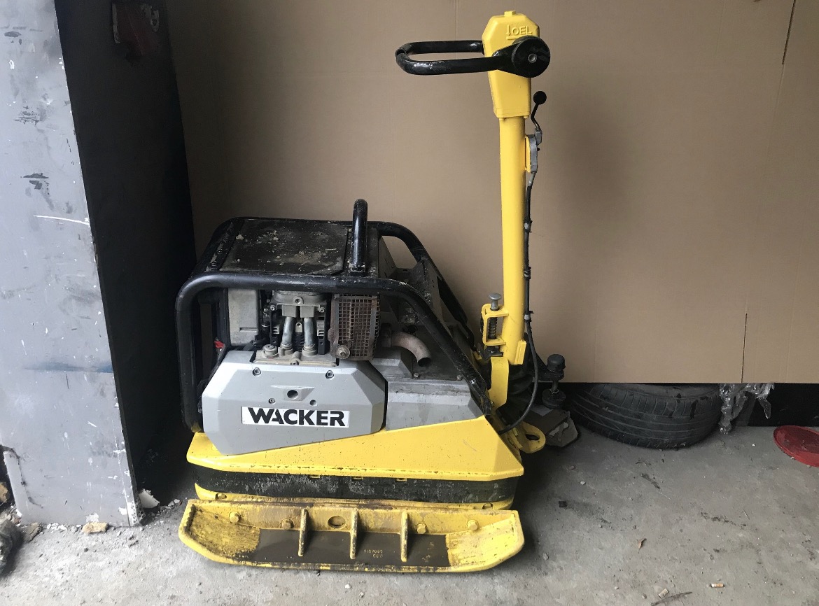 Żółto-szara zagęszczarka gruntu Wacker z czarną ramą i uchwytem, stojąca na betonowej powierzchni w pobliżu ściany, widoczne ślady użytkowania.