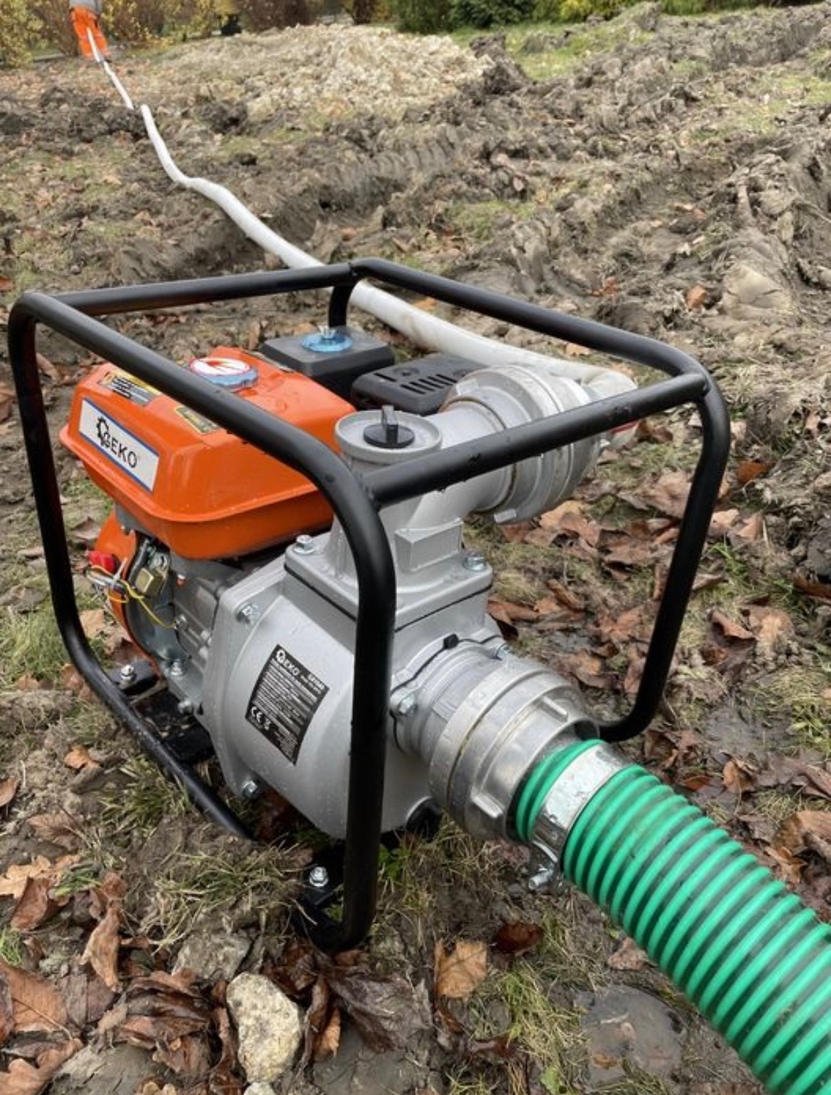Pompa spalinowa marki EKO z podłączonym zielonym wężem ssawnym, umieszczona na nierównym, błotnistym terenie z widocznymi śladami opon i białym wężem w tle.