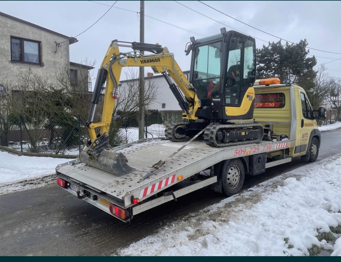 Żółty mini-koparka Yanmar transportowana na platformie lawety w zimowej scenerii; widoczny numer telefonu firmy transportowej.