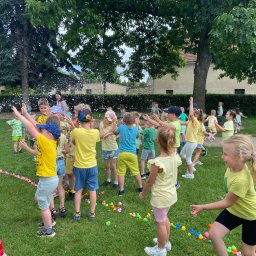 Magia Wrażeń Dominika Animator - Grupa dzieci w kolorowych ubraniach bawi się na trawie podczas imprezy plenerowej z animatorem, który puszcza sztuczny śnieg. W tle drzewa i budynki.