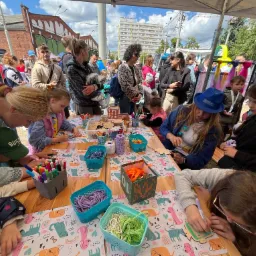 Warsztaty plastyczne dla dzieci: grupa dzieci przy stole dekoruje drewniane elementy, otoczone kolorowymi materiałami i pod nadzorem animatorów. Zabawa na świeżym powietrzu.