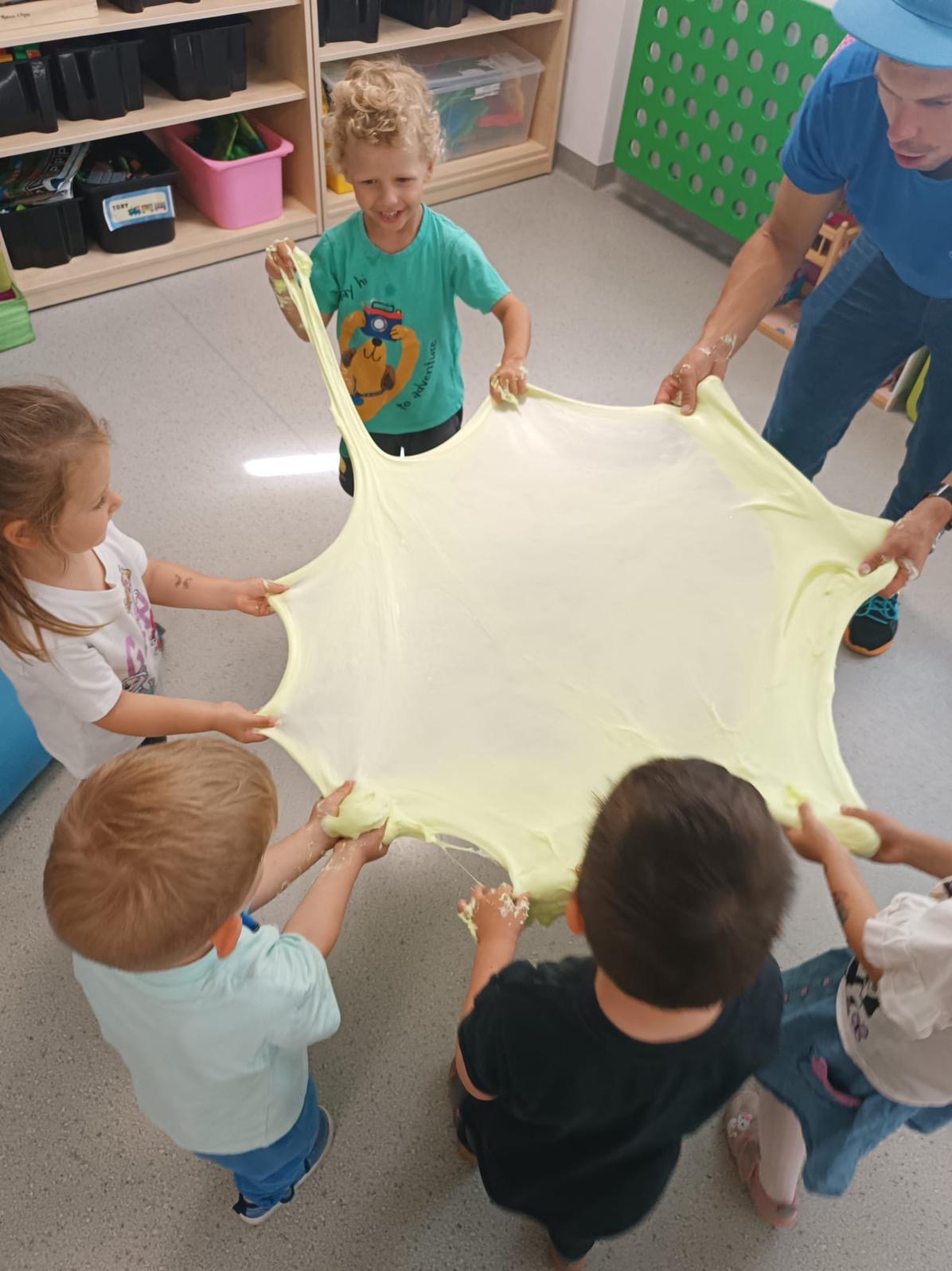 Dzieci z animatorem rozciągają dużą porcję żółtego slime'a w Raczycach. Zabawa angażuje grupę, w tle regał z zabawkami i jasna podłoga.