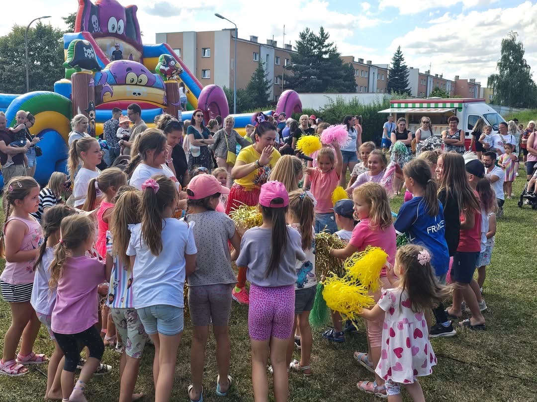 Animator w hawajskiej spódnicy prowadzi zabawę z pomponami dla grupy dzieci na tle dmuchanego zamku i tłumu ludzi.