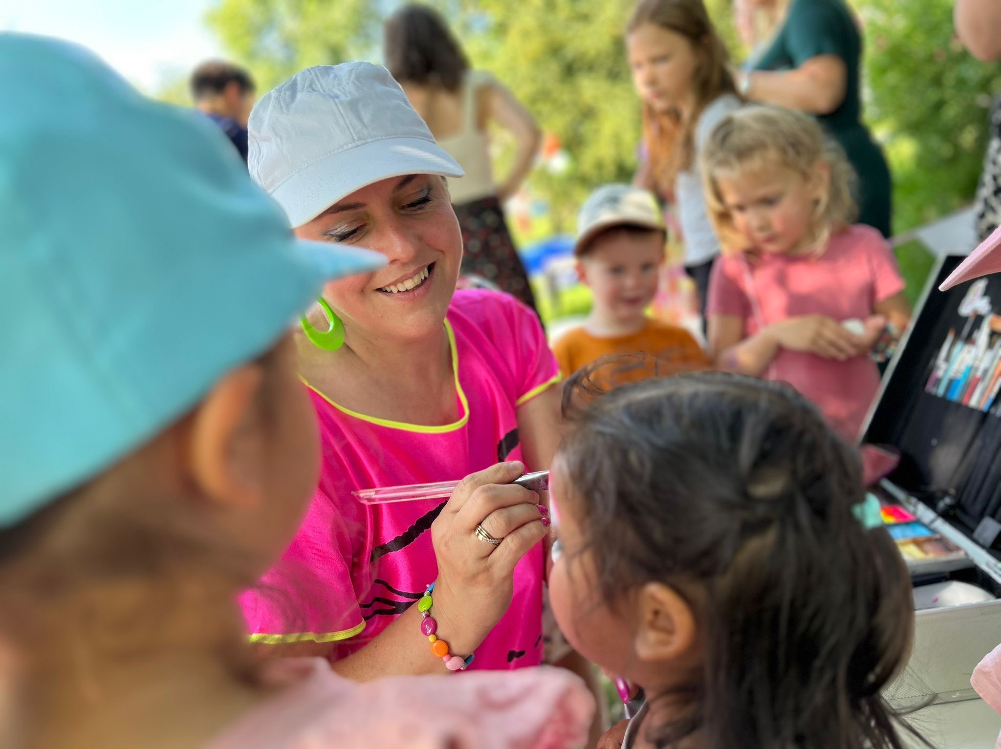 Animator maluje twarz dziewczynki pędzlem. W tle dzieci obserwują, a na stole widoczny zestaw farb. Kobieta w różowej bluzce i białej czapce uśmiecha się.