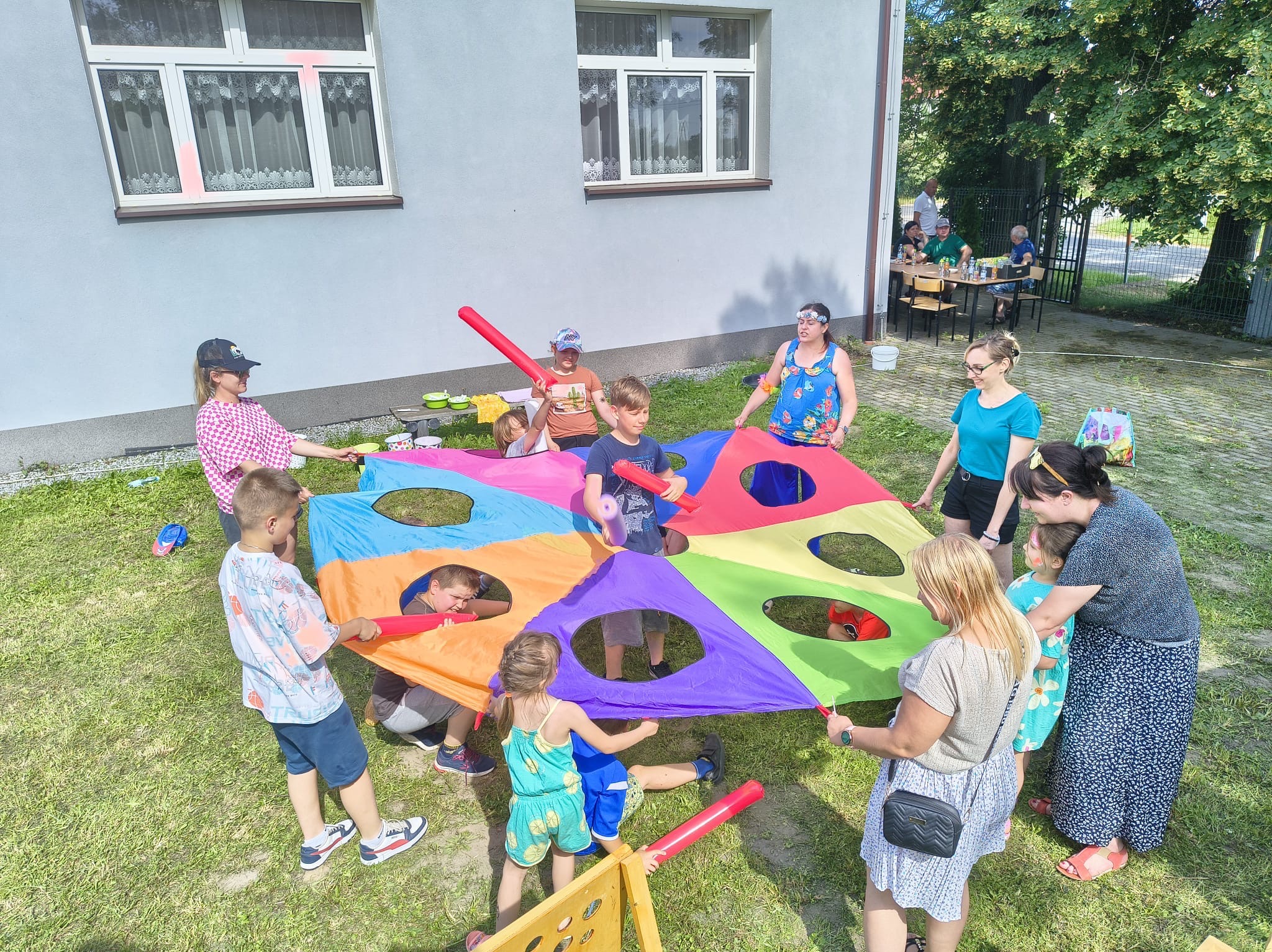 Grupa dzieci i dorosłych bawi się kolorową chustą animacyjną na trawie podczas imprezy plenerowej. W tle budynek i osoby siedzące przy stole.