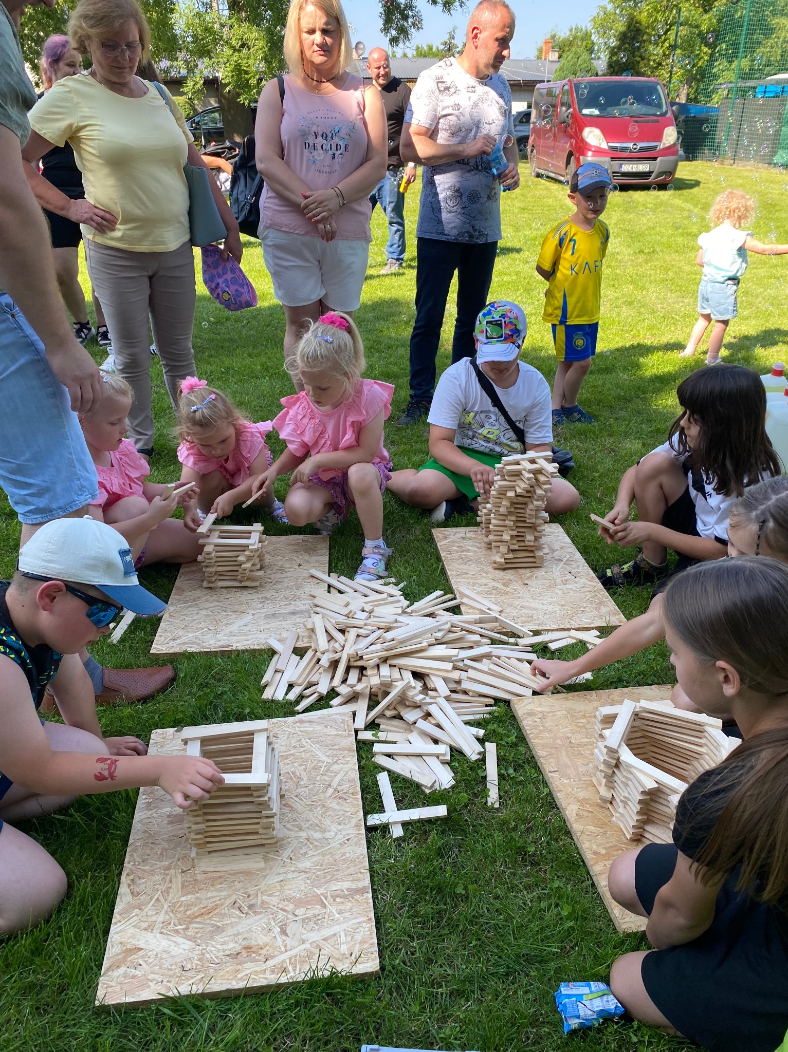 Dzieci na trawie budują konstrukcje z drewnianych klocków na deskach podczas animacji na świeżym powietrzu. W tle rodzice i czerwony van.