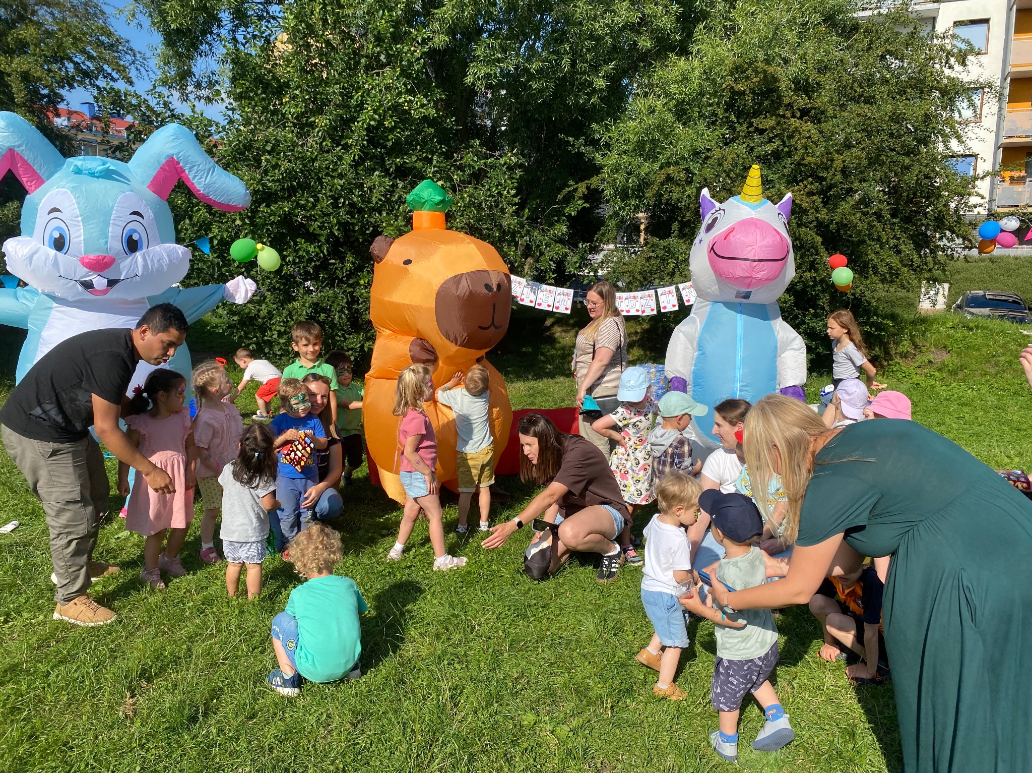 Animatorzy w akcji: dmuchany królik, miś i jednorożec otoczeni grupą dzieci podczas imprezy plenerowej na trawie, z opiekunami w tle.