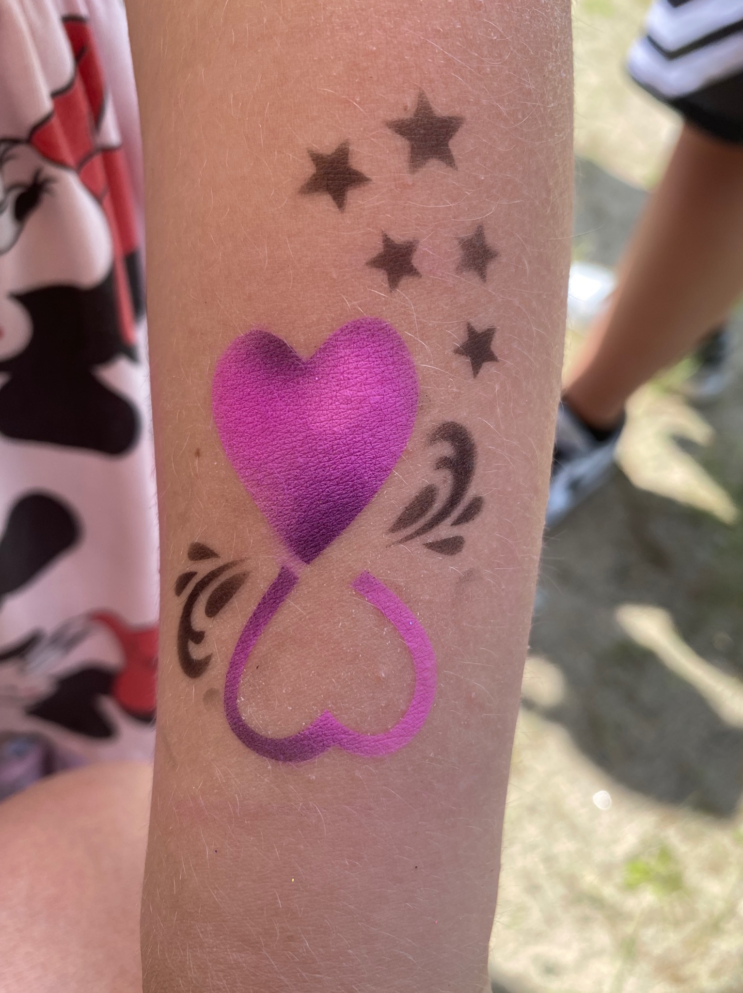 Dziecięce ramię z fioletowym tatuażem: dwa serca połączone w klepsydrę, czarne gwiazdki i zawijasy. Jasne tło, widoczny fragment ubrania dziecka.