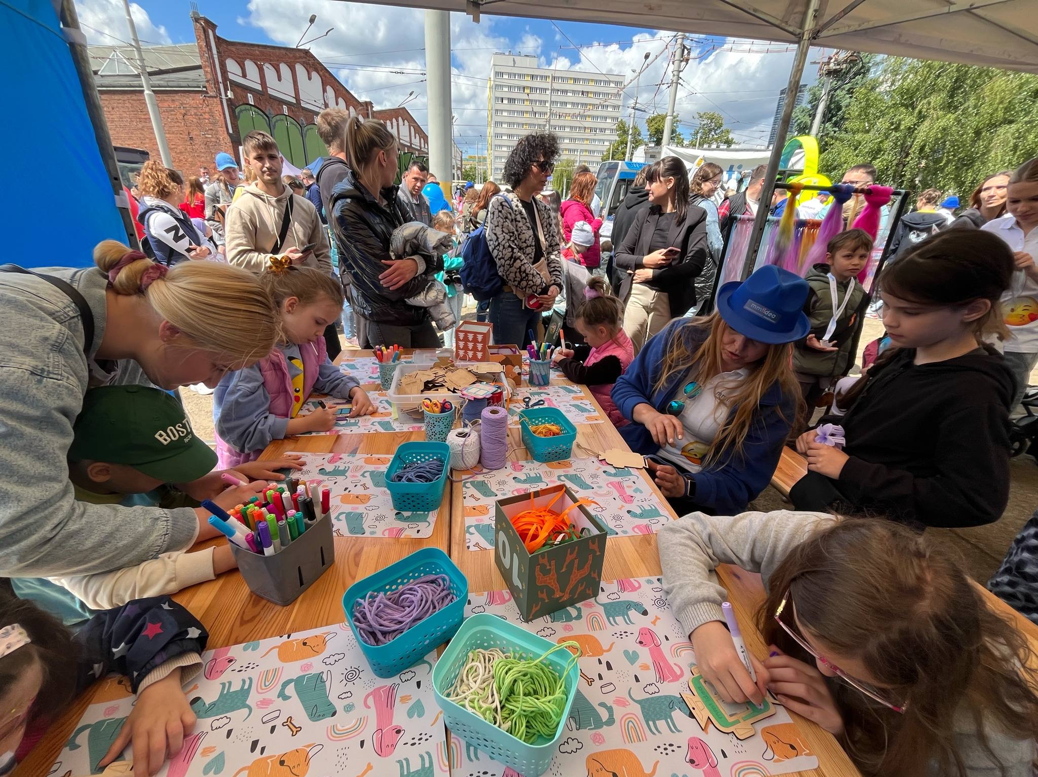 Warsztaty plastyczne dla dzieci: grupa dzieci przy stole dekoruje drewniane elementy, otoczone kolorowymi materiałami i pod nadzorem animatorów. Zabawa na świeżym powietrzu.