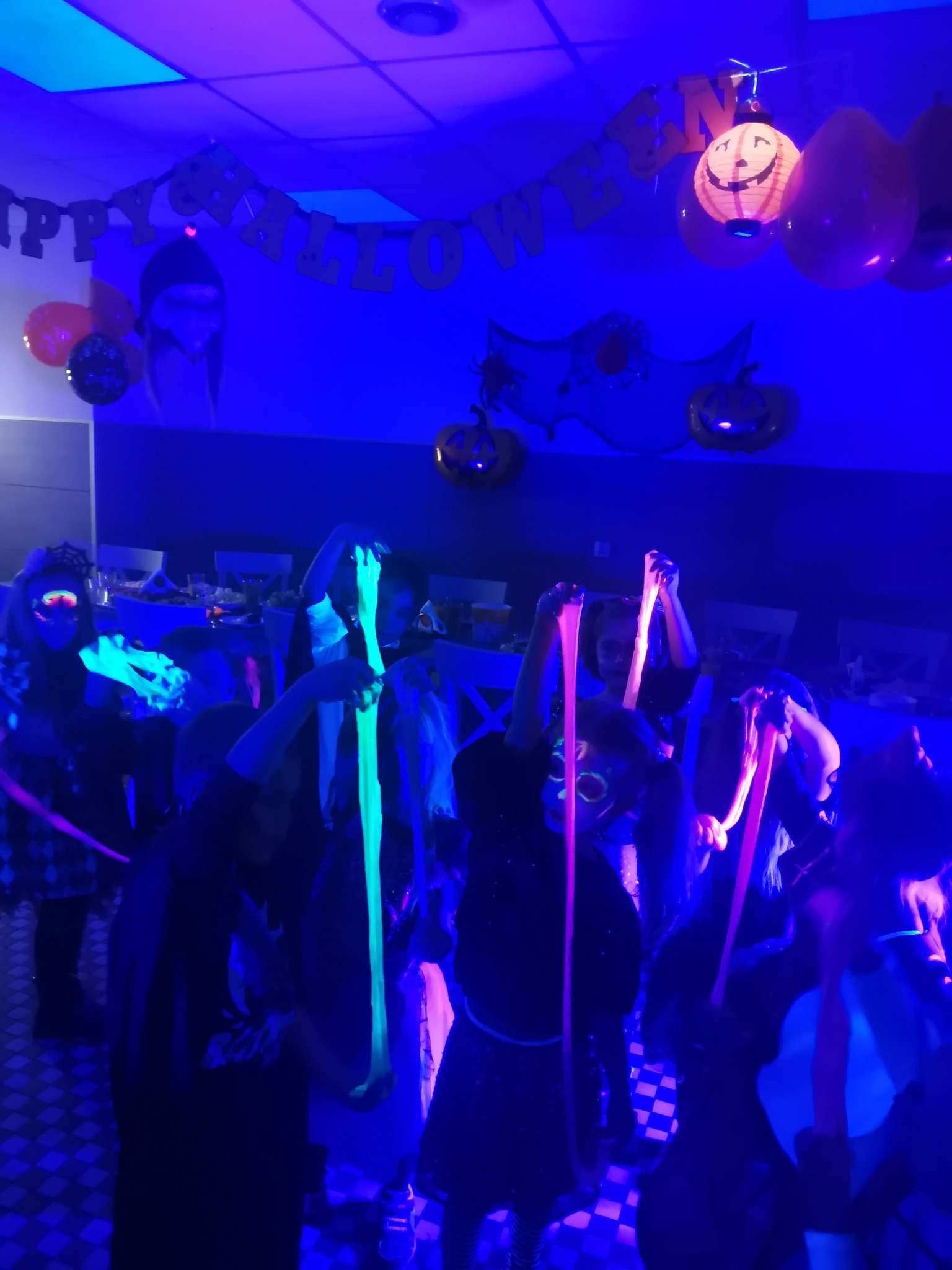 Dzieci z pomalowanymi twarzami i świecącymi w ciemności slime bawią się na imprezie Halloween, dekoracje z dyniami i napisem Happy Halloween podświetlone niebieskim światłem UV.