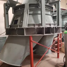 Szara, przemysłowa konstrukcja stalowa z rurami i łańcuchami, wsparta na czerwonym stelażu, widoczna w hali produkcyjnej.