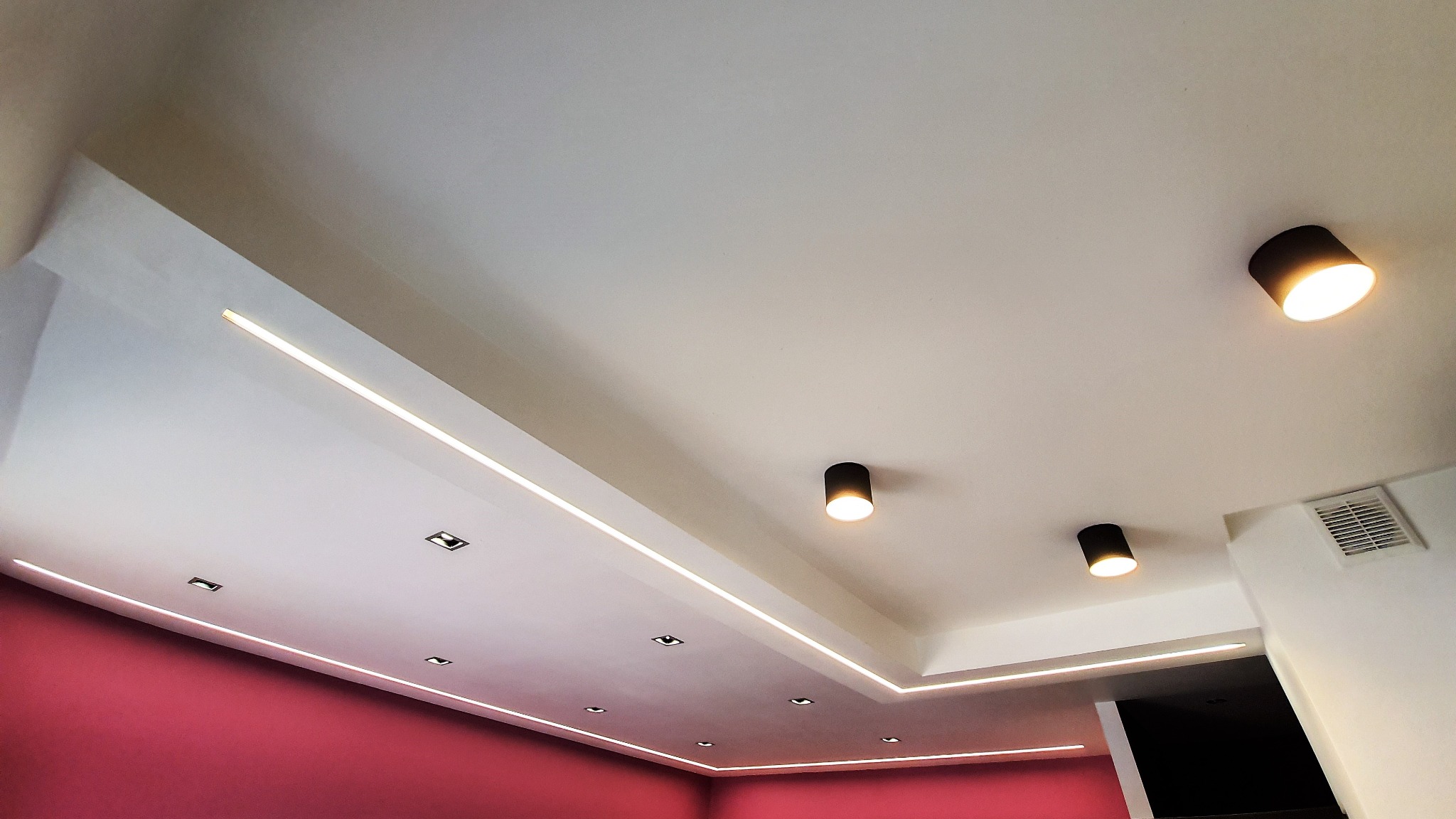 Nowoczesne oświetlenie sufitowe z liniowymi lampami LED i punktowymi reflektorami w kolorze czarnym, ściana pomalowana na różowo.