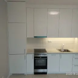 Biała zabudowa kuchenna z płytkami imitującymi cegłę na ścianie, widoczny zlewozmywak, płyta grzewcza i piekarnik Amica.