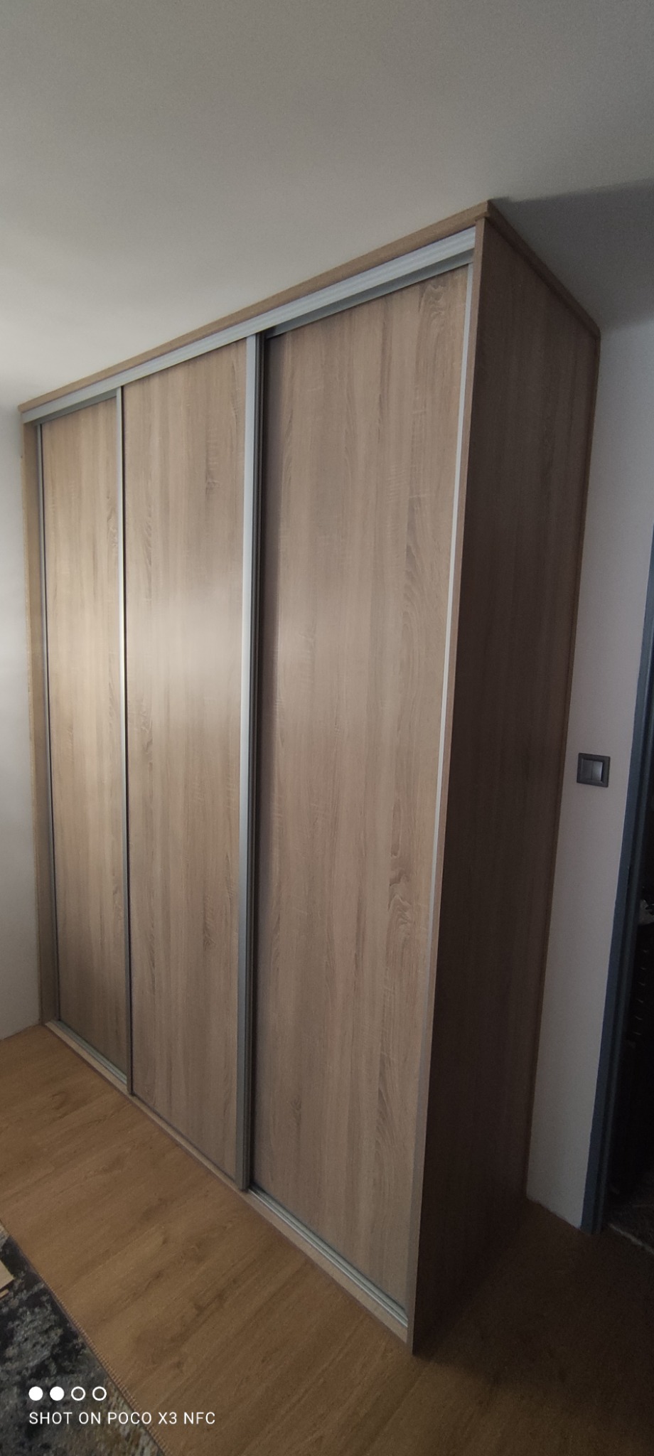 Trzydrzwiowa szafa przesuwna z frontami w kolorze jasnego drewna dębowego, z srebrnymi prowadnicami, ustawiona na podłodze z paneli, widok z boku.