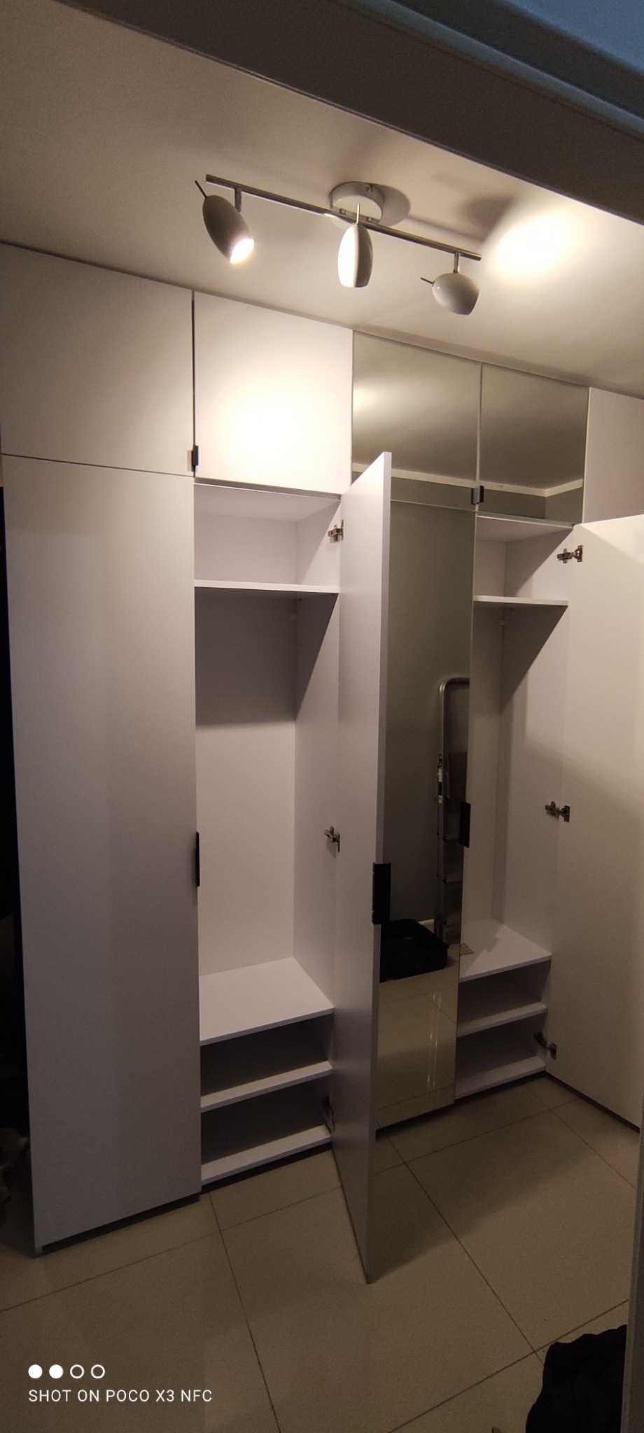 Biała szafa wnękowa z lustrzanymi drzwiami, oświetleniem punktowym i otwartymi półkami, uchwycona w kadrze z perspektywy podłogi.
