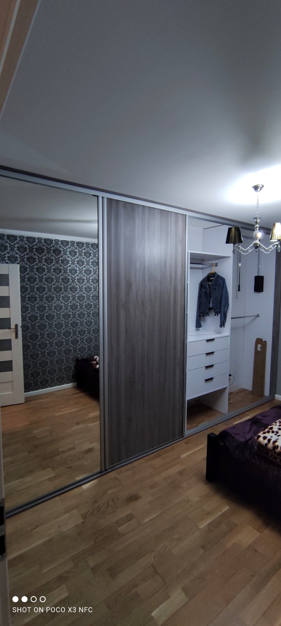 Nowoczesna szafa z przesuwnymi drzwiami, z lustrem i drewnopodobnym wykończeniem, w sypialni z parkietem i wzorzystą tapetą, w tle widoczna garderoba z szufladami i wiszącą kurtką jeansową.