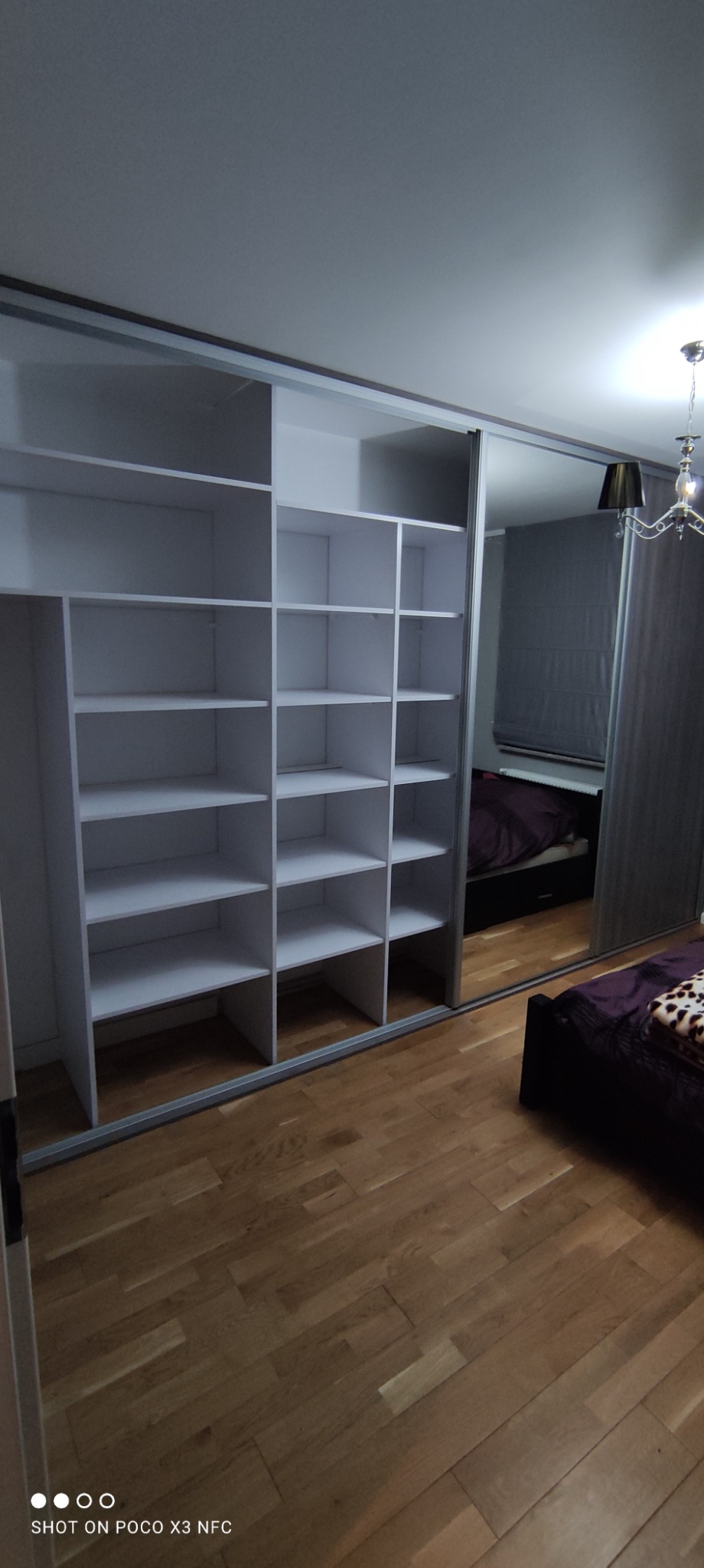 Biała, wnękowa szafa z przesuwnymi drzwiami lustrzanymi, widoczna na tle sypialni z drewnianą podłogą.