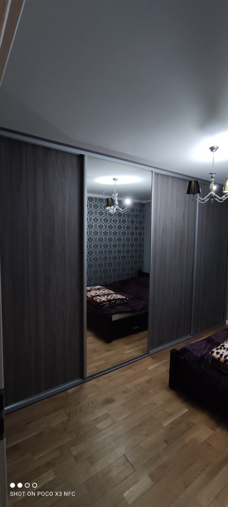 Szara szafa przesuwna z lustrem, odbijająca sypialnię z łóżkiem i wzorzystą tapetą, parkiet na podłodze.