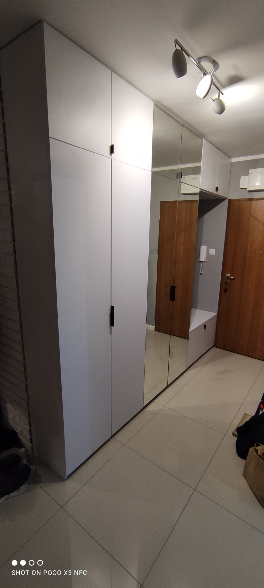 Biała szafa wnękowa z lustrzanymi drzwiami, czarnymi uchwytami, z siedziskiem i szafką przy wejściu do mieszkania.