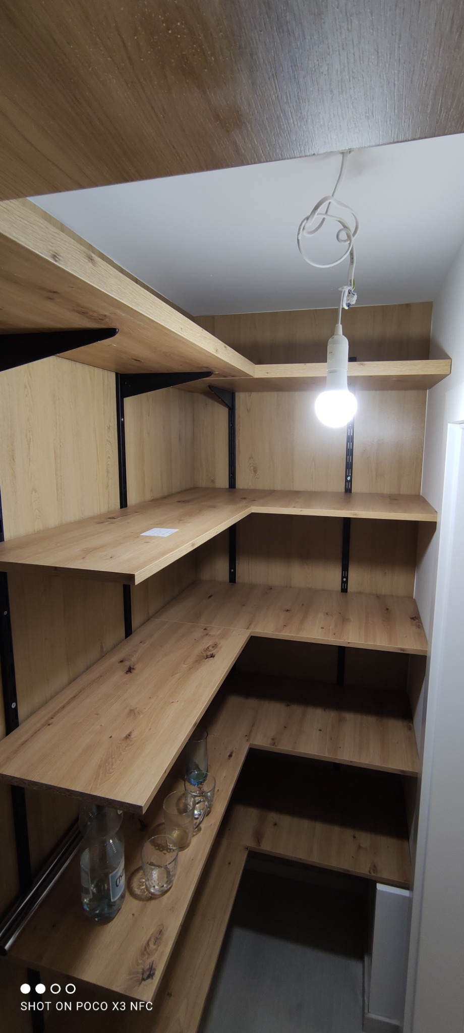 Wnętrze spiżarni z półkami z płyty w odcieniach drewna, wspartych na czarnych, metalowych wspornikach, oświetlone wiszącą żarówką; na dolnej półce szklane naczynia i butelka.