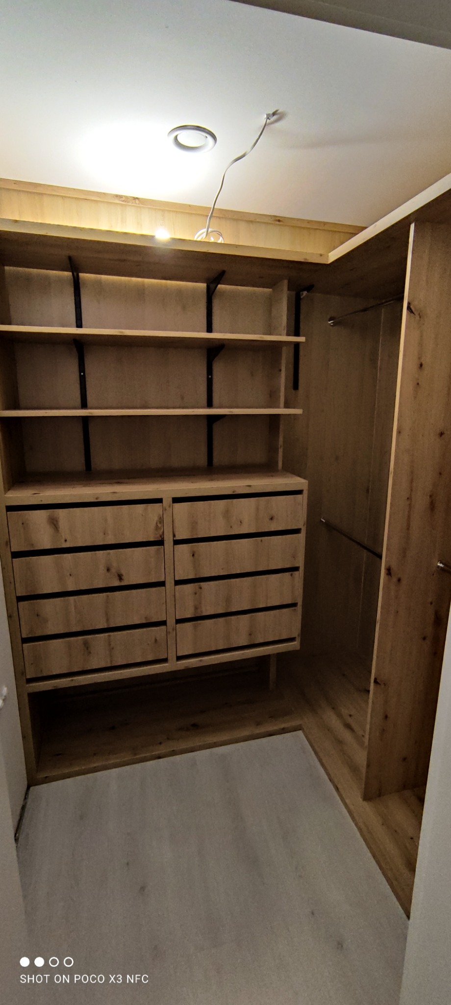 Wnętrze garderoby z drewnianymi półkami na czarnych wspornikach i szufladami z ciemnymi uchwytami, widok z dołu na sufit z wystającym kablem oświetleniowym.