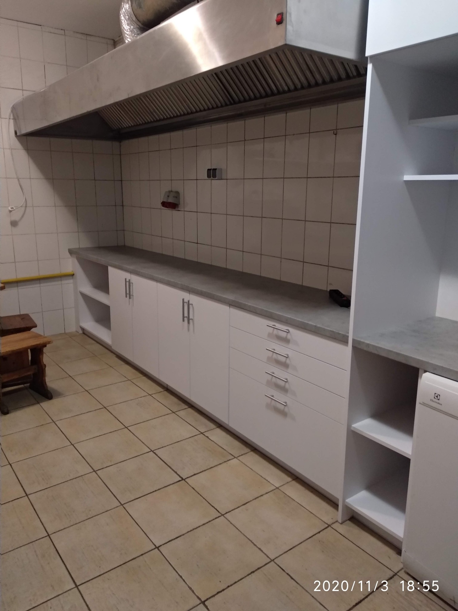 Wnętrze kuchni z białymi szafkami z szarym blatem, widoczny fragment okapu i biała lodówka Electrolux po prawej stronie.