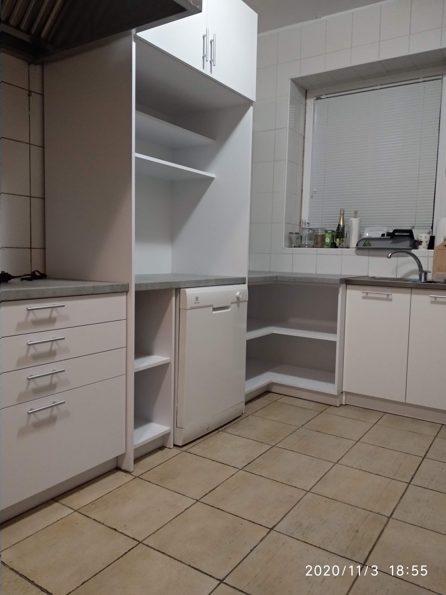 Biała zabudowa kuchenna z szufladami, szafkami i półkami, zintegrowana ze sprzętem AGD, na tle białych płytek ściennych i jasnej podłogi.