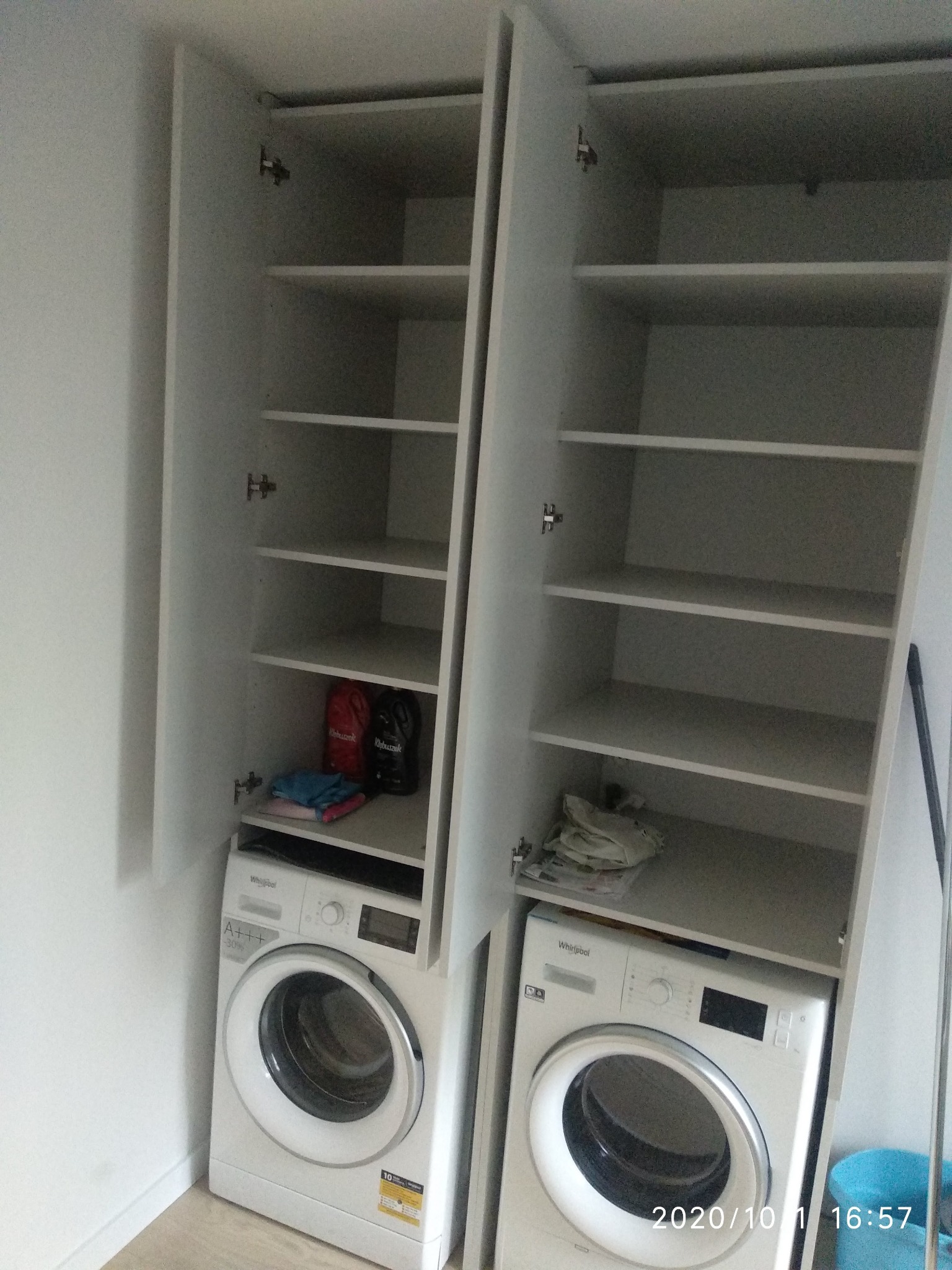 Dwie białe pralki umieszczone w szafkach z otwartymi drzwiami, widoczne półki i detergenty na dolnych półkach.