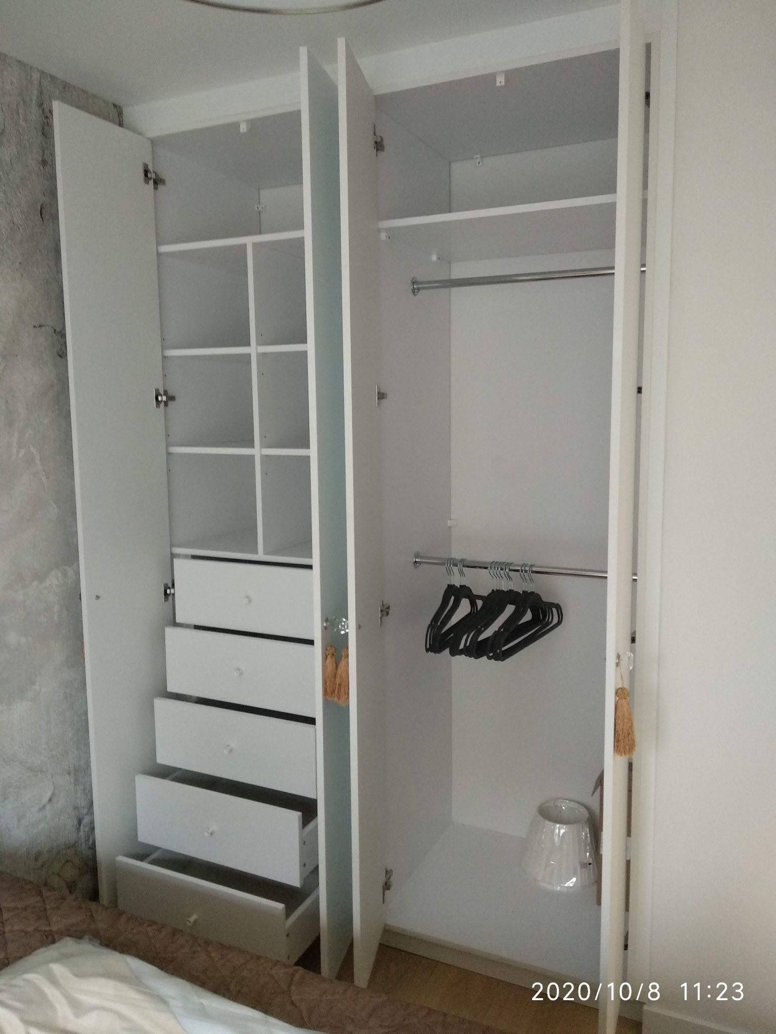 Wnętrze białej szafy wnękowej z otwartymi drzwiami, widoczne półki, szuflady z kryształowymi gałkami i drążek na wieszaki z czarnymi wieszakami, na podłodze lampa z abażurem w folii.