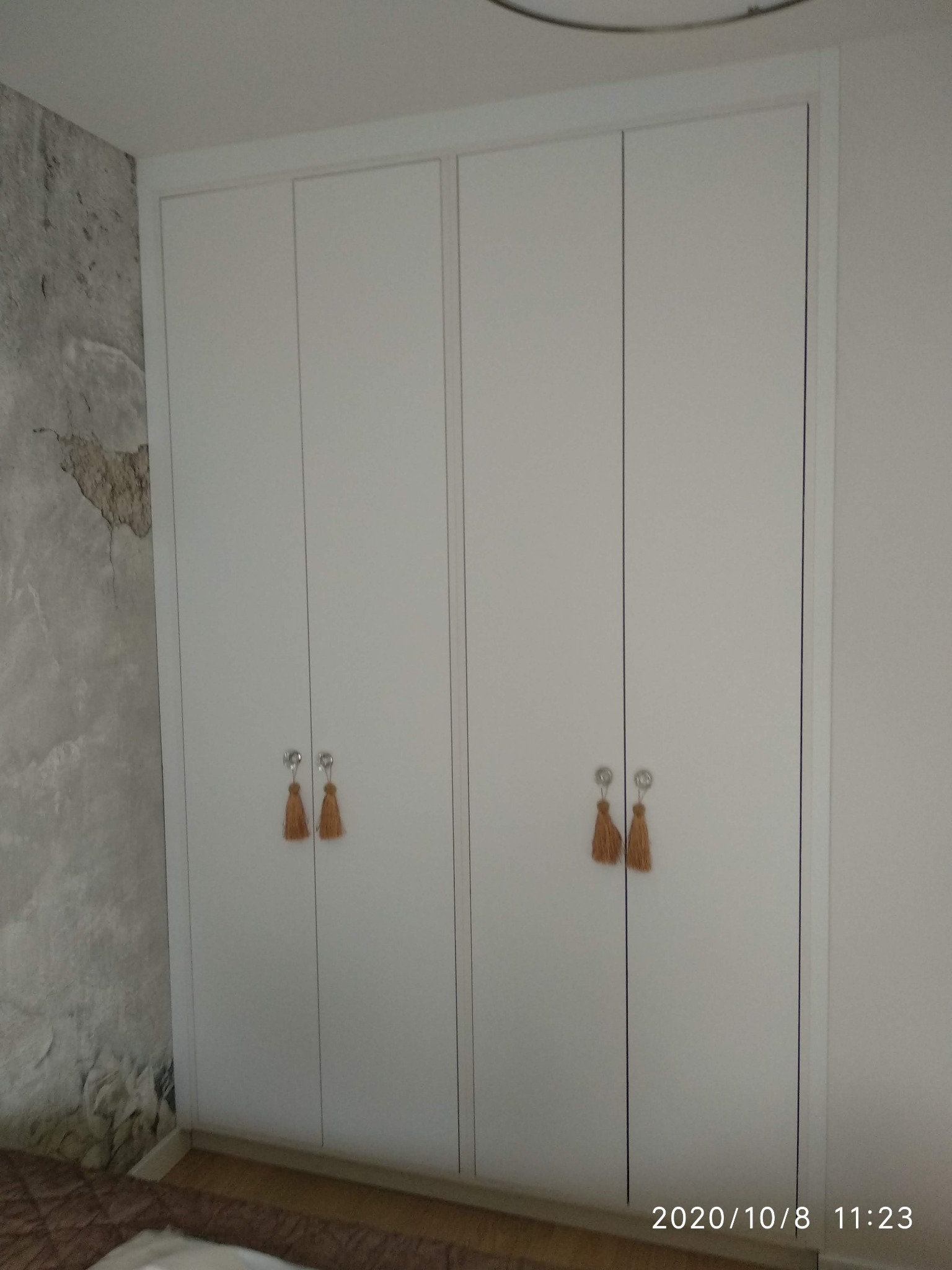 Biała, czterodrzwiowa szafa z delikatnymi uchwytami z brązowymi frędzlami, wkomponowana w pokój z dekoracyjną ścianą imitującą surowy beton.